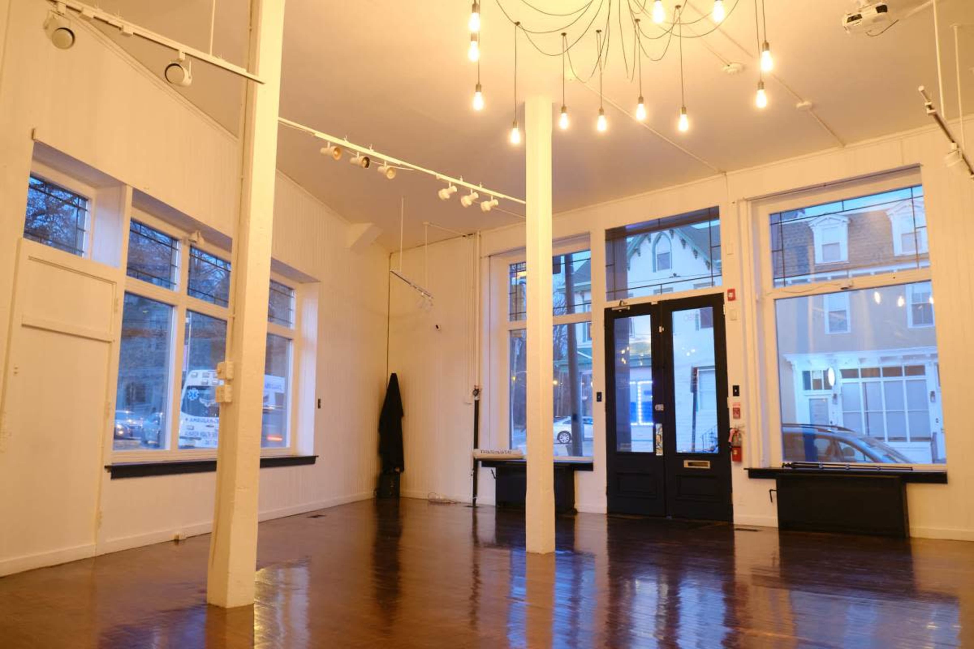 The image shows an empty interior space with wooden floors, large windows, and several hanging light fixtures.