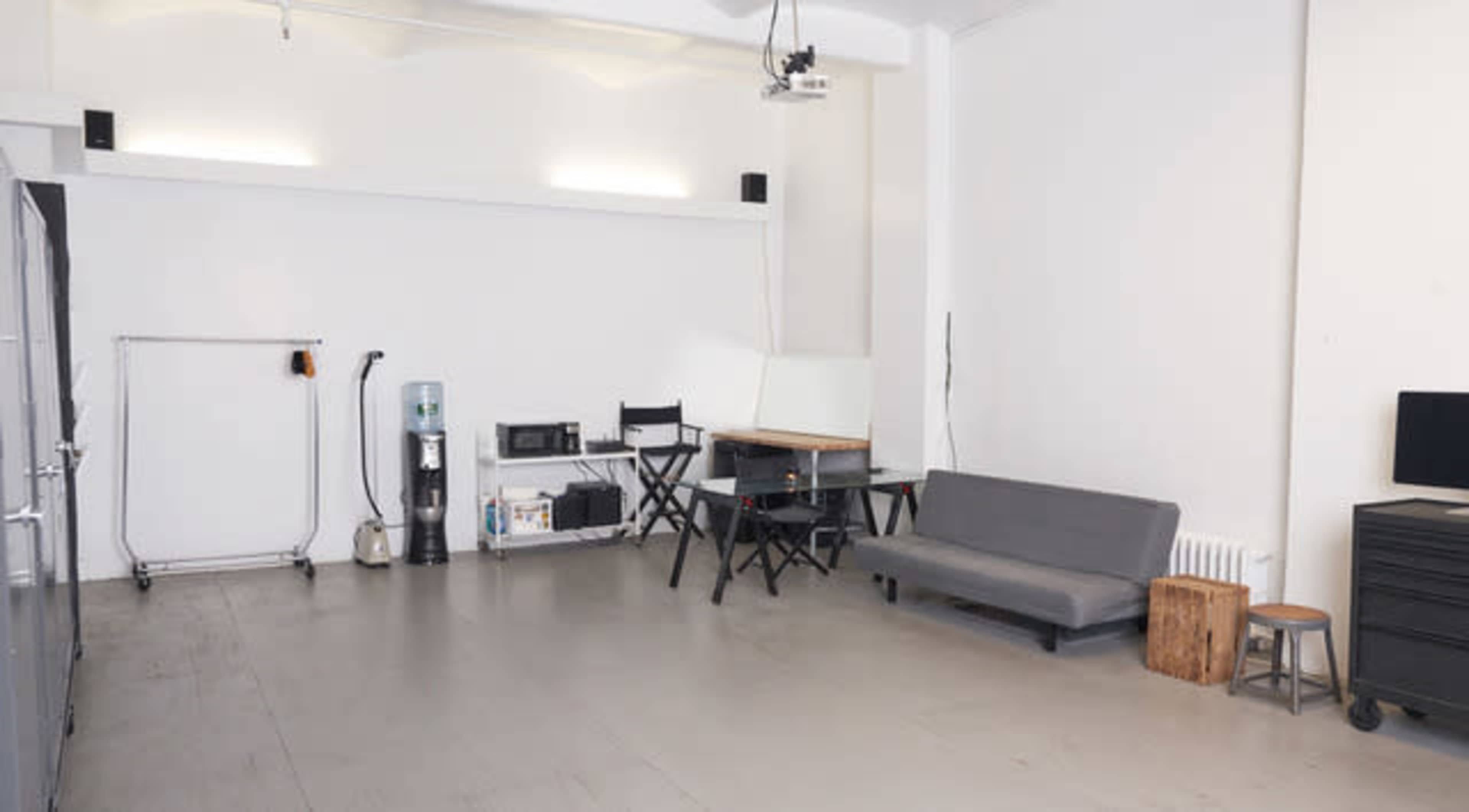 The image shows a spacious, minimalist room featuring a gray sofa, a wooden stool, a table with chairs, a water cooler, and various appliances against a plain white wall.