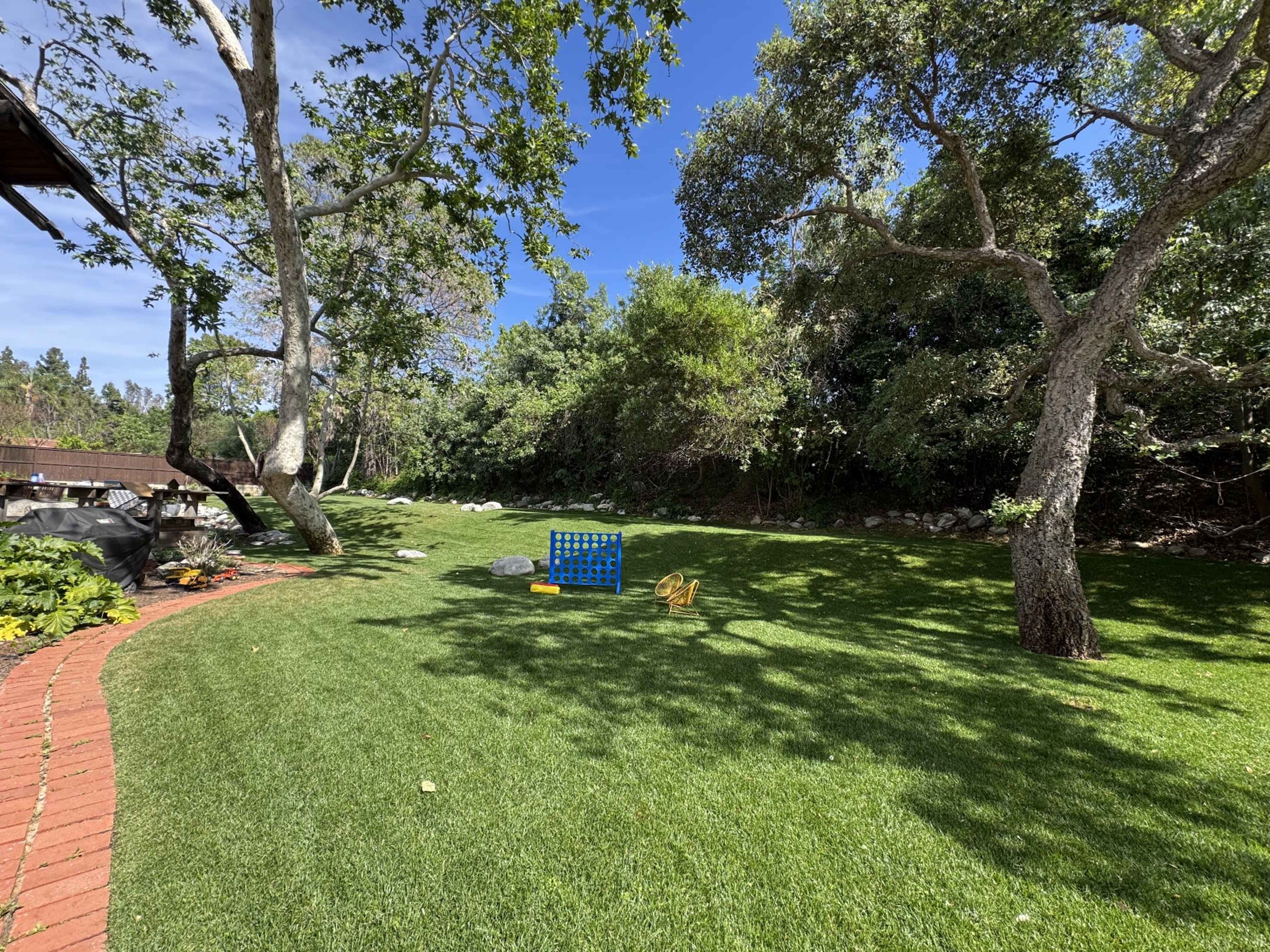 The scene features a grassy backyard with a curved brick path, large trees, and outdoor games set up, including a Connect Four game.