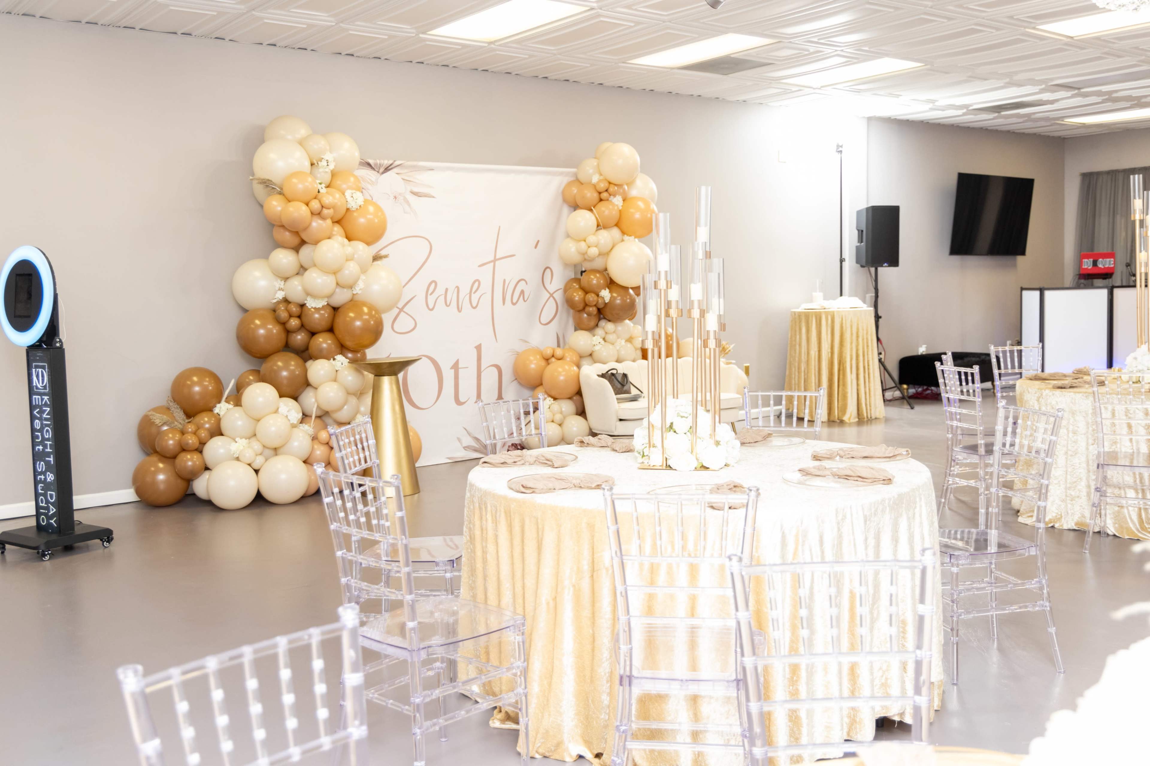 The image shows a decorated event space featuring elegant round tables with golden tablecloths, clear chairs, and a backdrop of balloons in shades of gold and beige.
