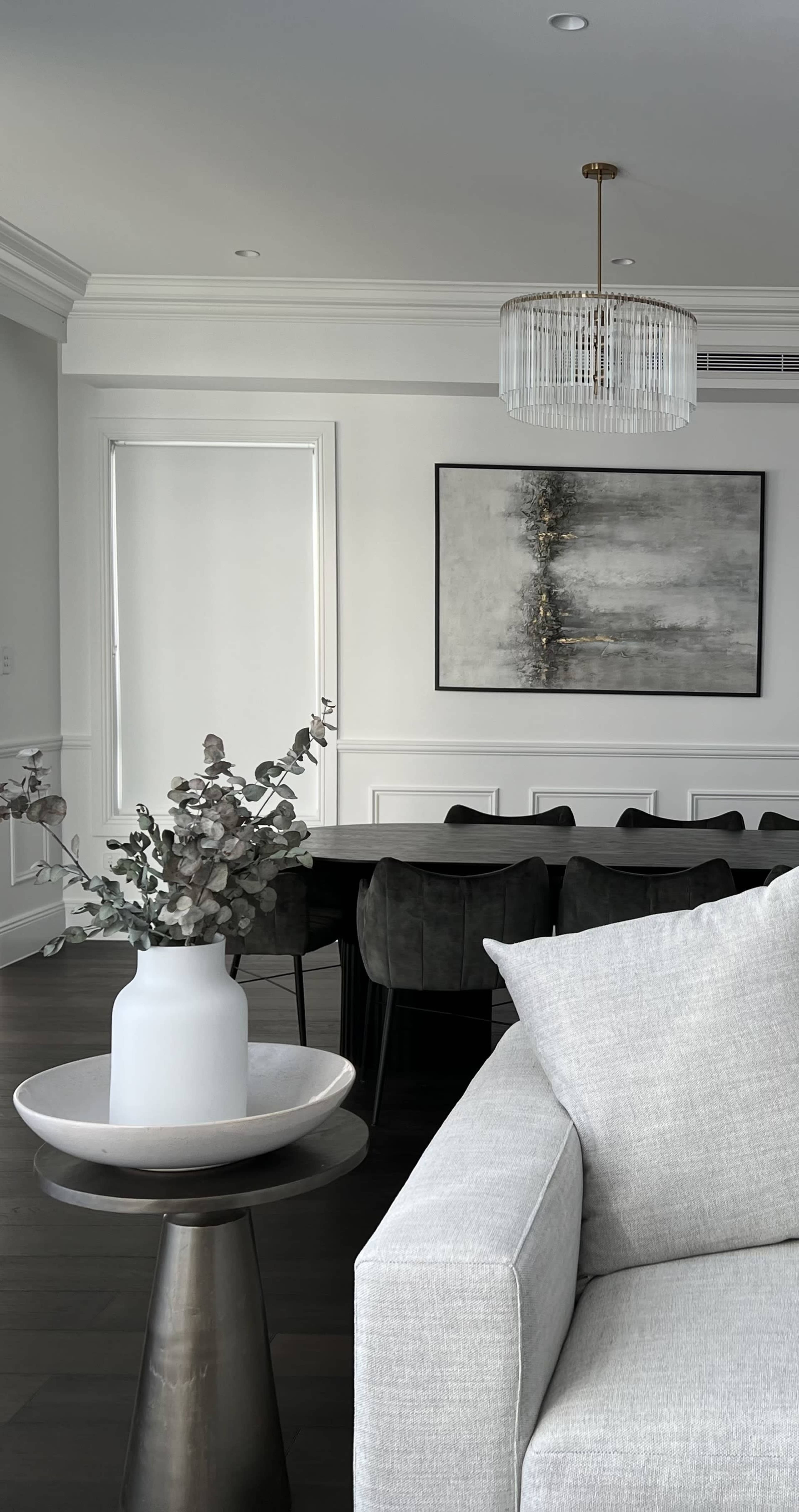A modern living room features a light gray sofa, a decorative table with a white vase and plant, and a dining area with a dark table and chairs, set against a background of light walls and artwork.