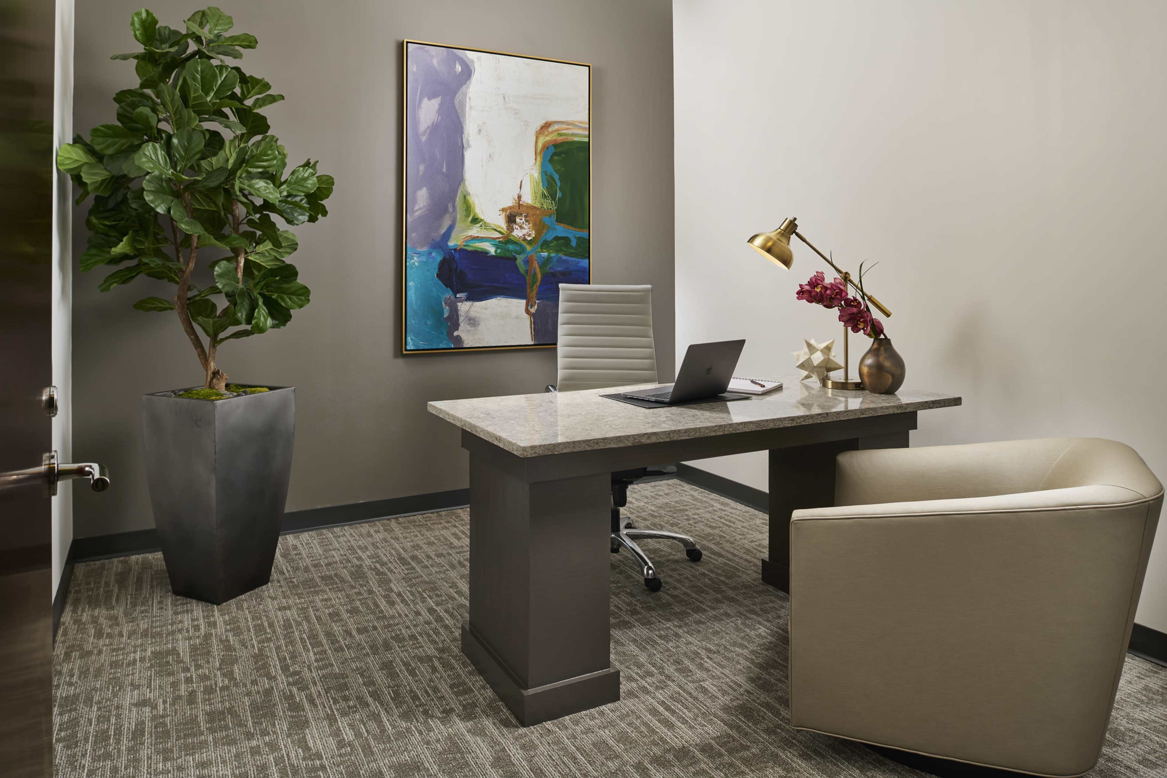 An office features a sleek desk with a laptop, a comfortable chair, a potted plant, and a large piece of artwork on the wall.