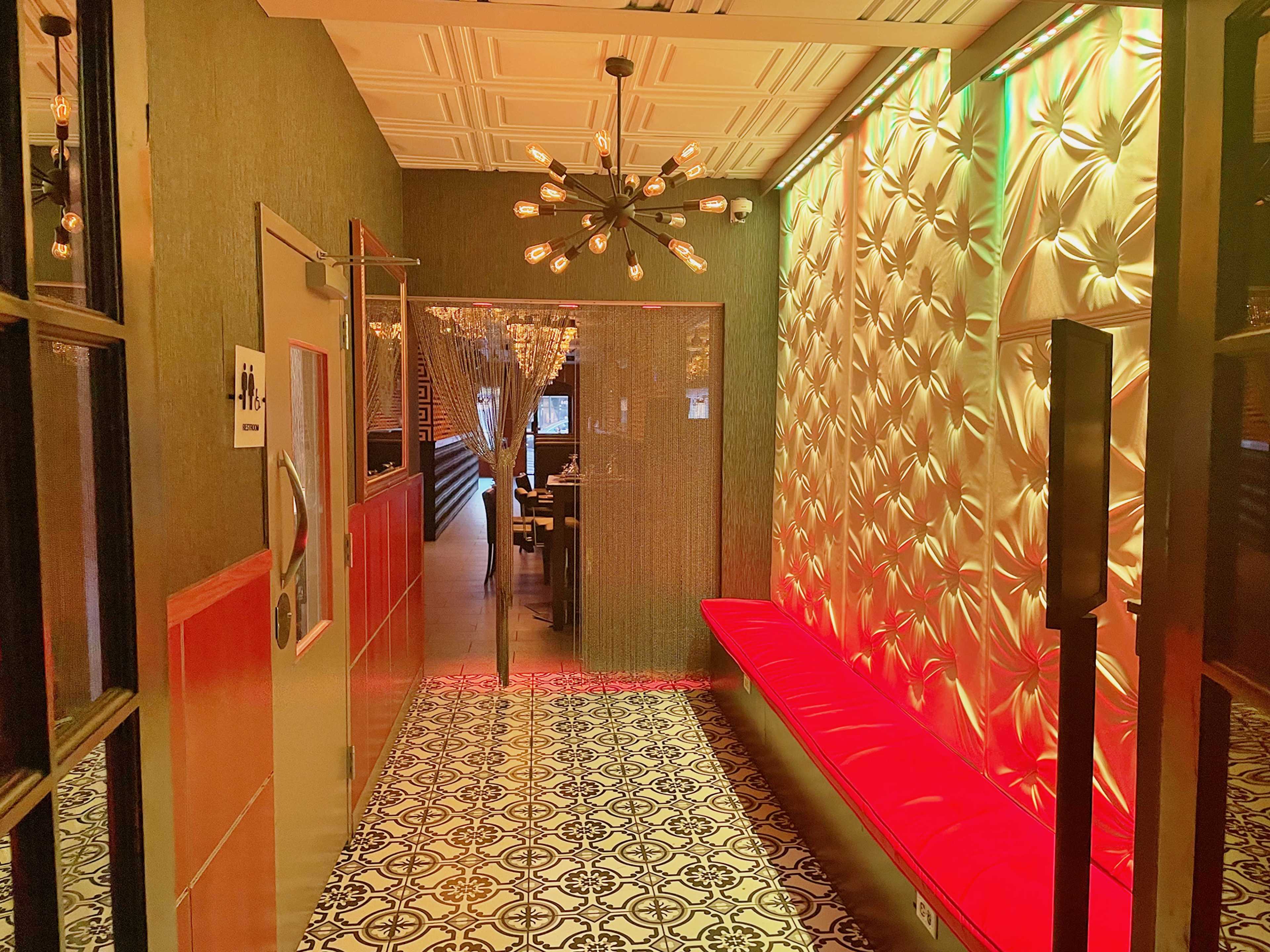 A narrow hallway leading to a dining area, featuring a decorative wall, a red bench, patterned flooring, and stylish lighting.