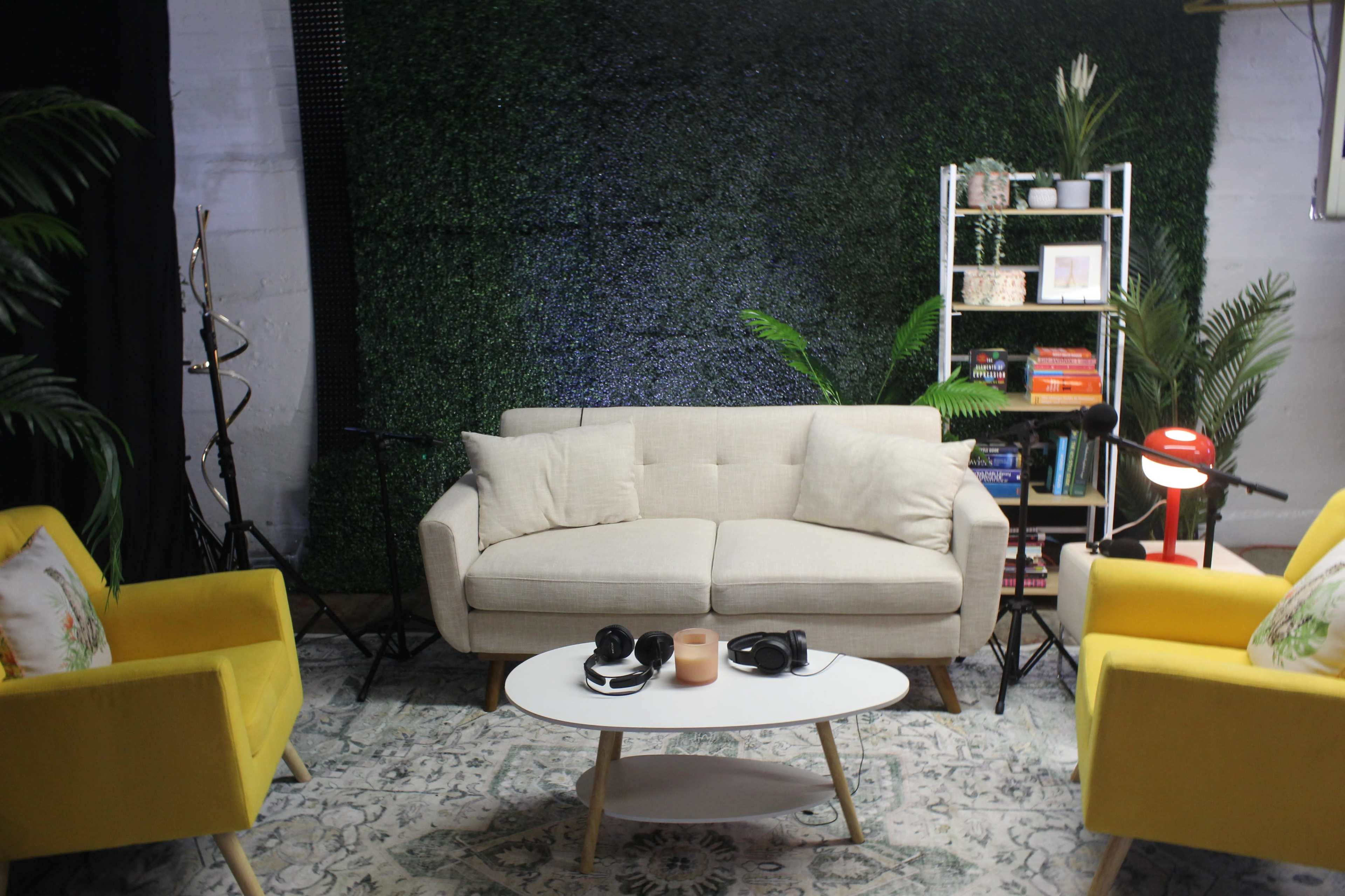 A modern seating area features a light-colored sofa and two yellow armchairs around a white coffee table, set against a green wall of artificial plants.