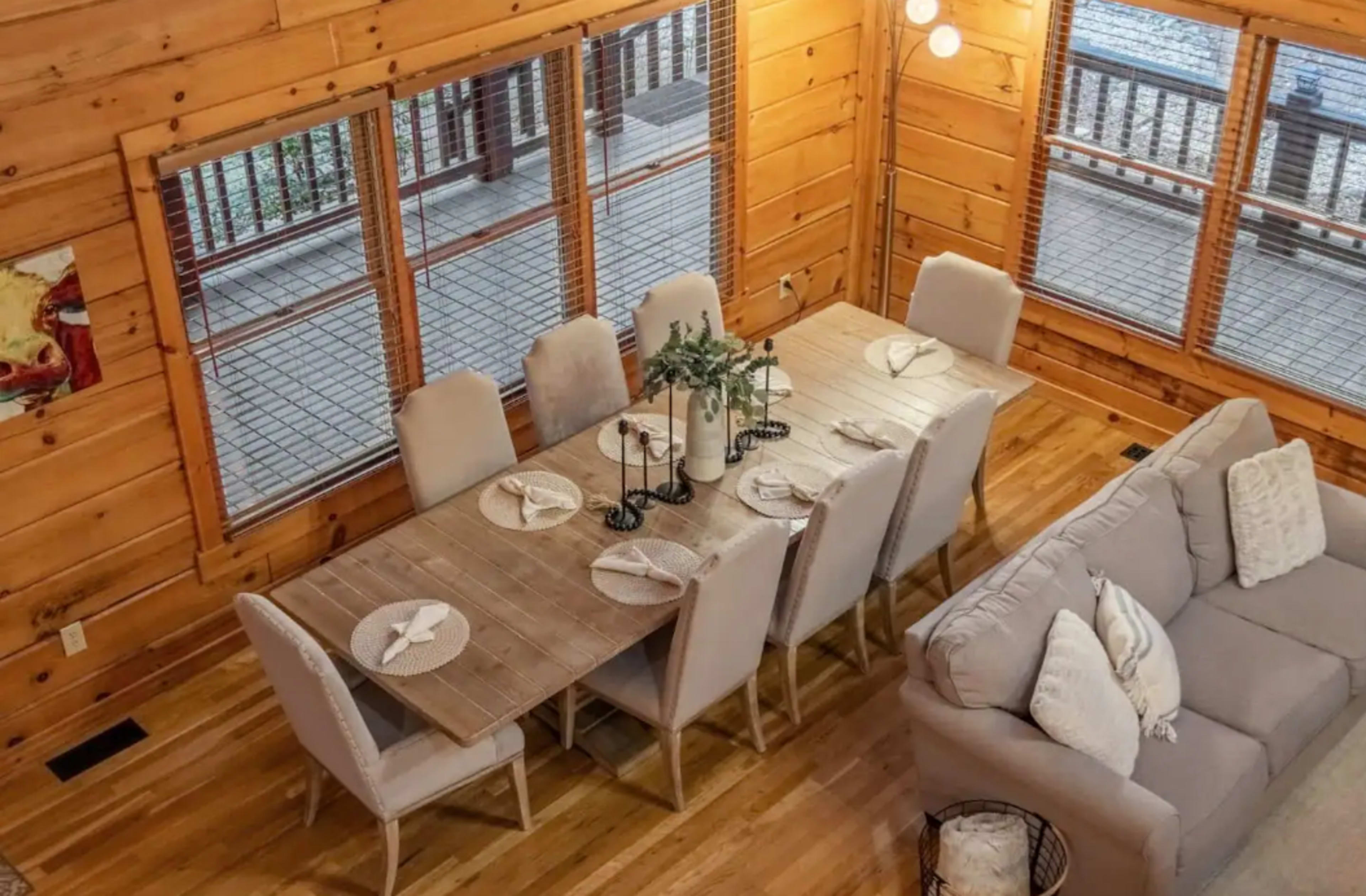 A wooden dining area features a long table set for eight, surrounded by upholstered chairs, with large windows providing outside views.