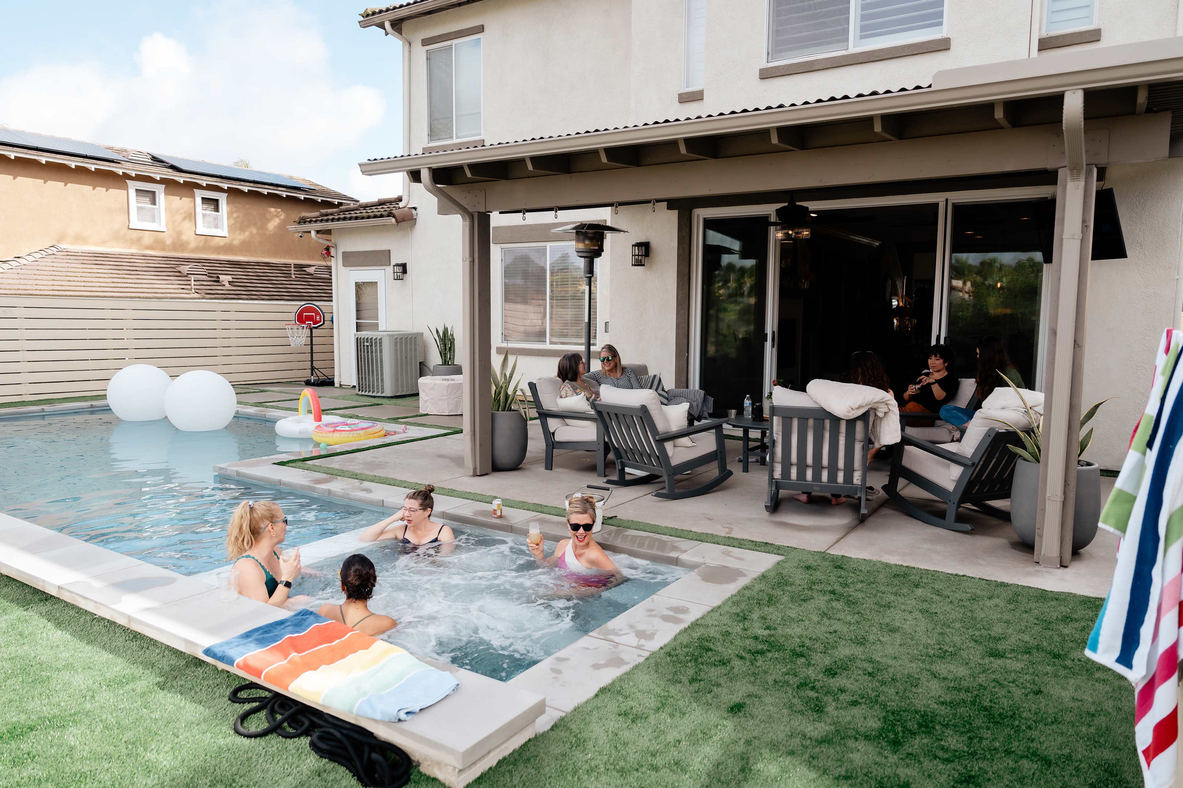 A group of people socializes by a poolside while some relax in a hot tub, with lounge chairs and a patio area visible in the background.