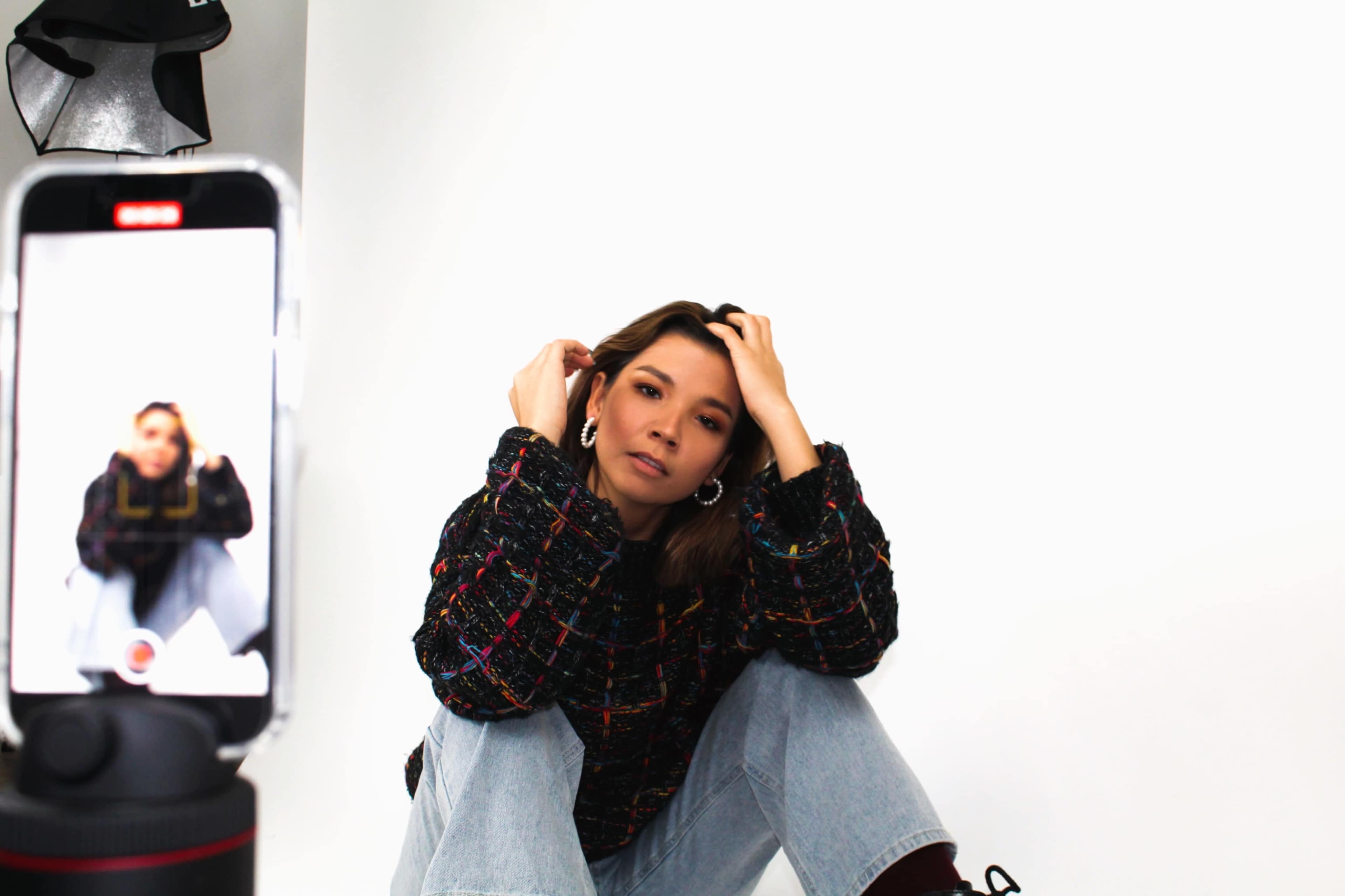 A woman in a colorful sweater sits on the floor while posing for a photo taken with a smartphone.