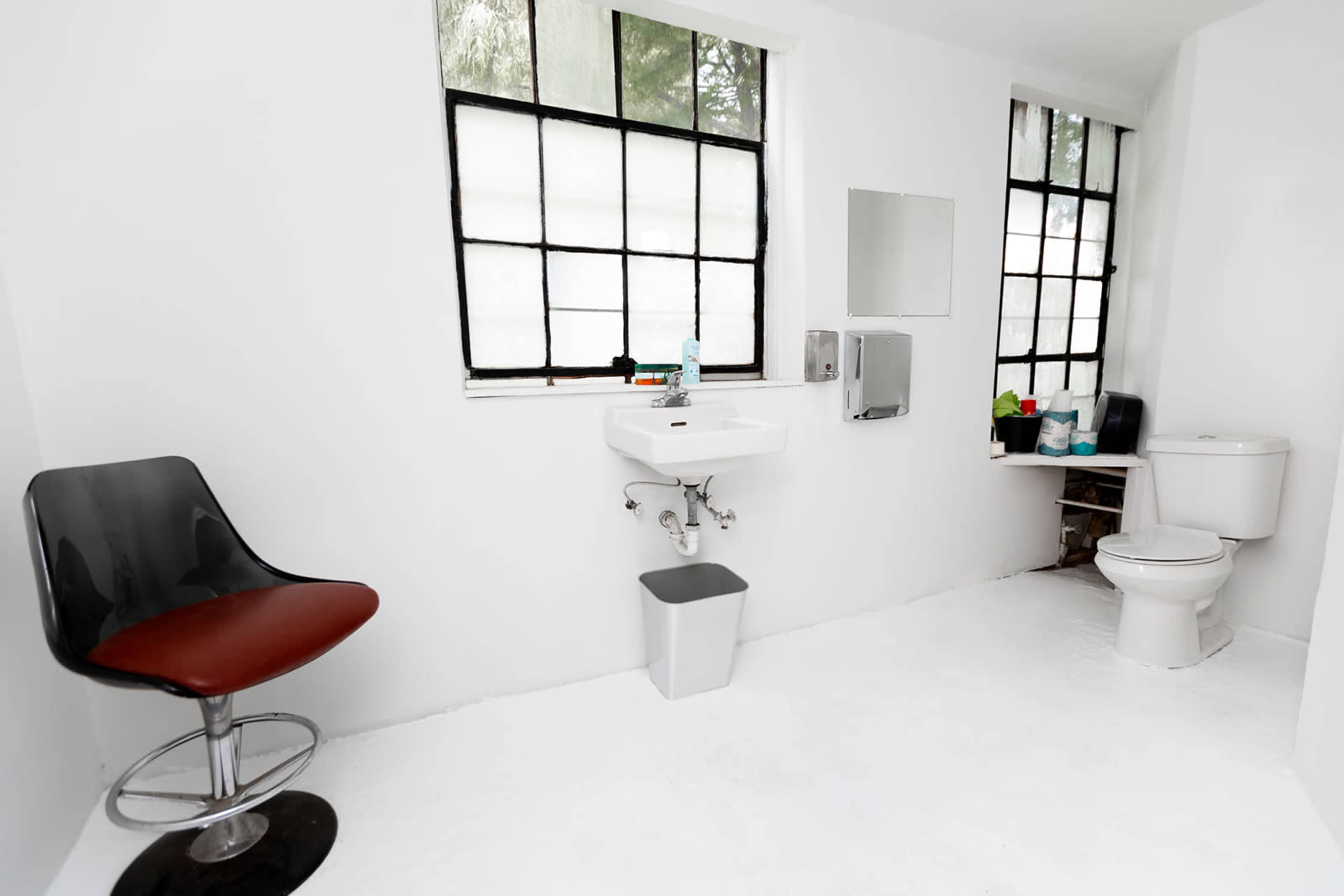 The image shows a small bathroom with a toilet, sink, a mirror, and a modern chair positioned near a window.