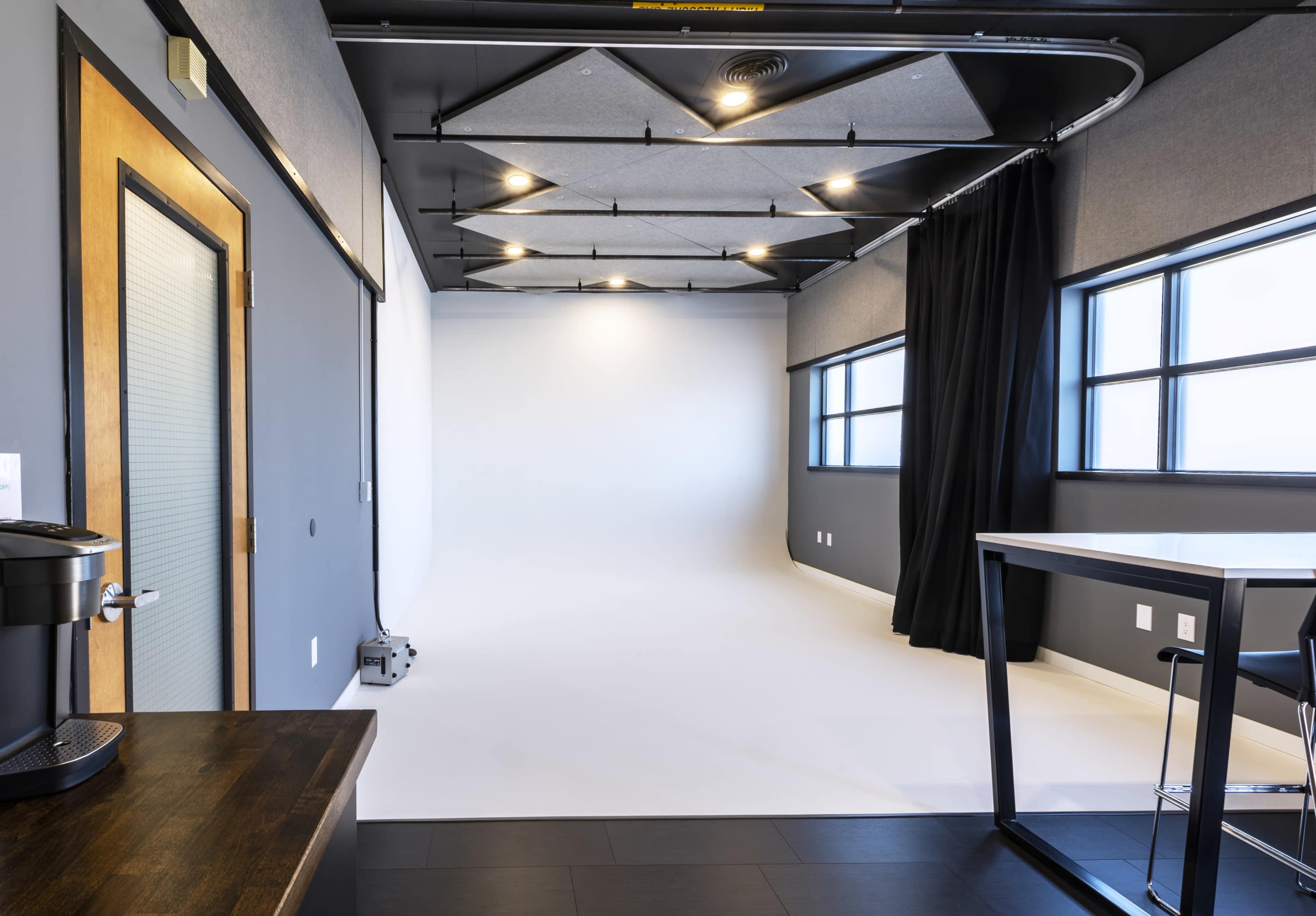 A modern studio space with a white backdrop, large windows, a coffee station, and a black curtain.