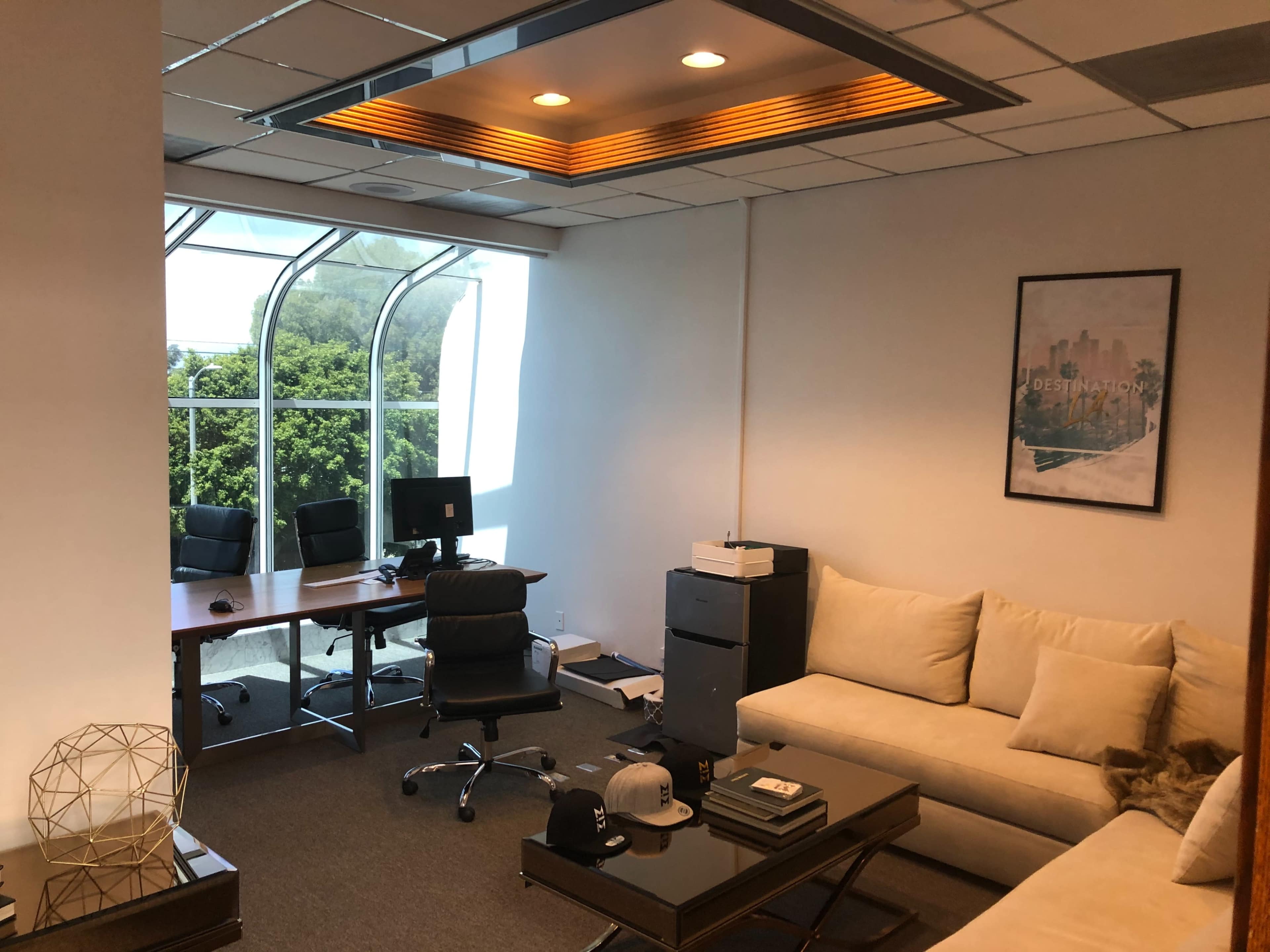 An office space featuring a large window, a sectional sofa, a coffee table, and a workspace with desks and office equipment.