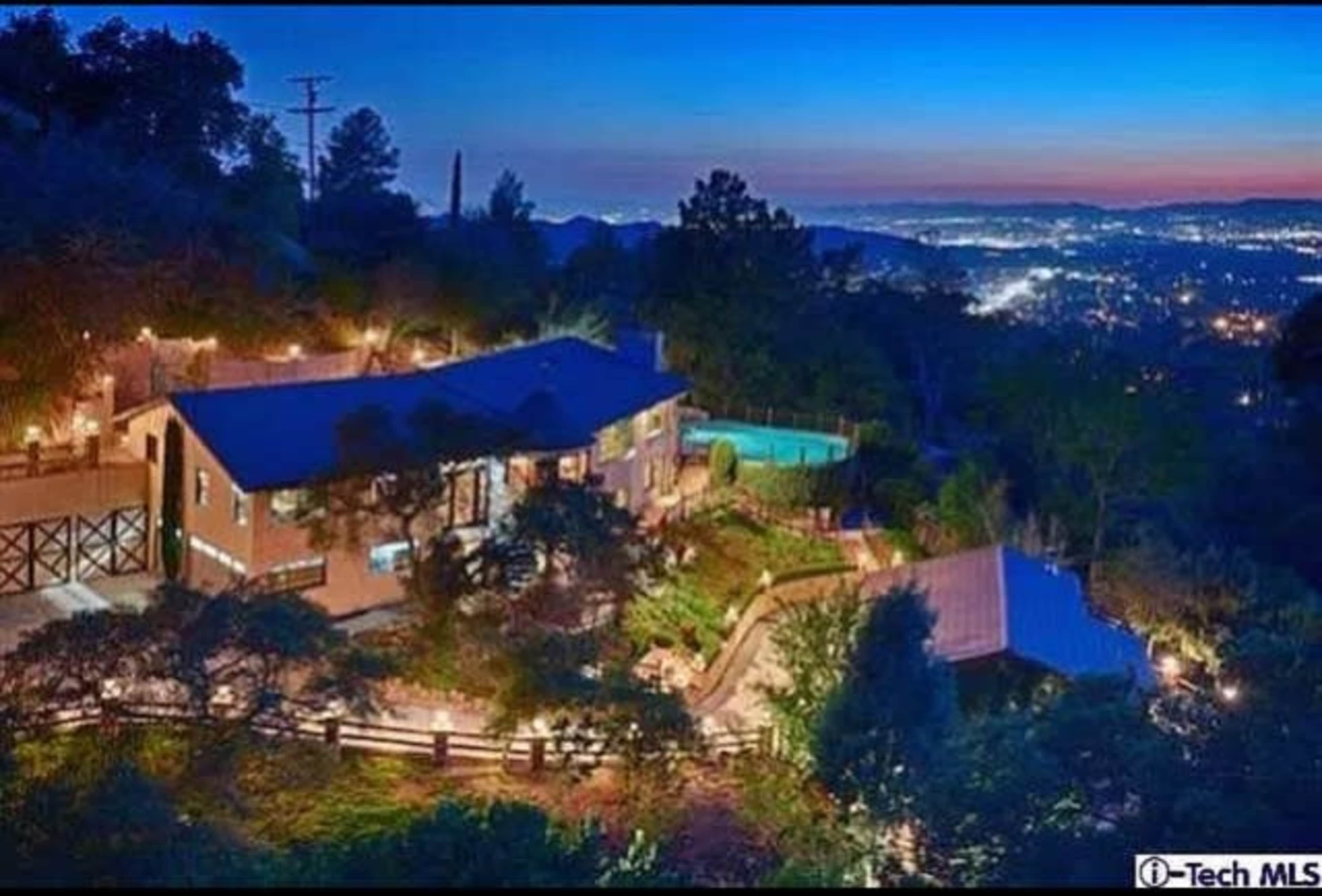 The image shows a hillside home illuminated at night, featuring a swimming pool and a panoramic view of the city below.