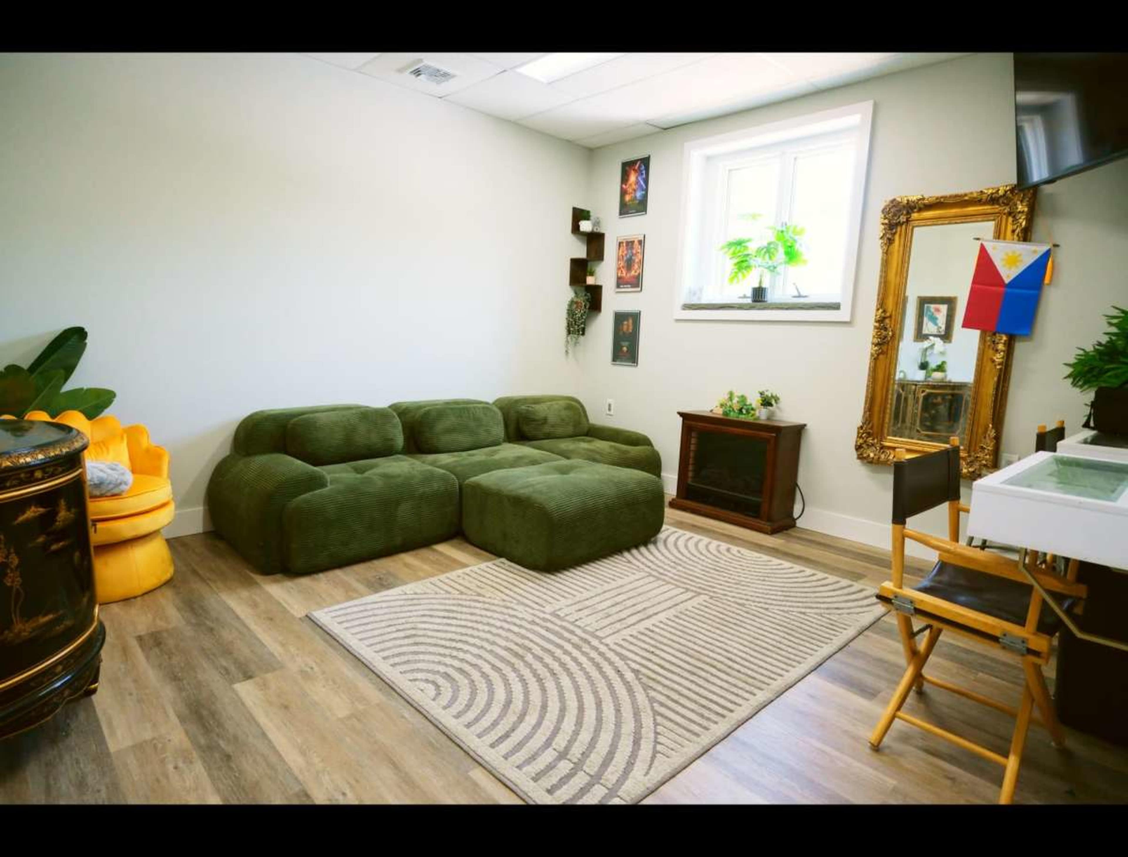 The image depicts a tidy room featuring a green modular sofa, a wooden cabinet, a decorative mirror, a small table with a chair, and various wall decorations.