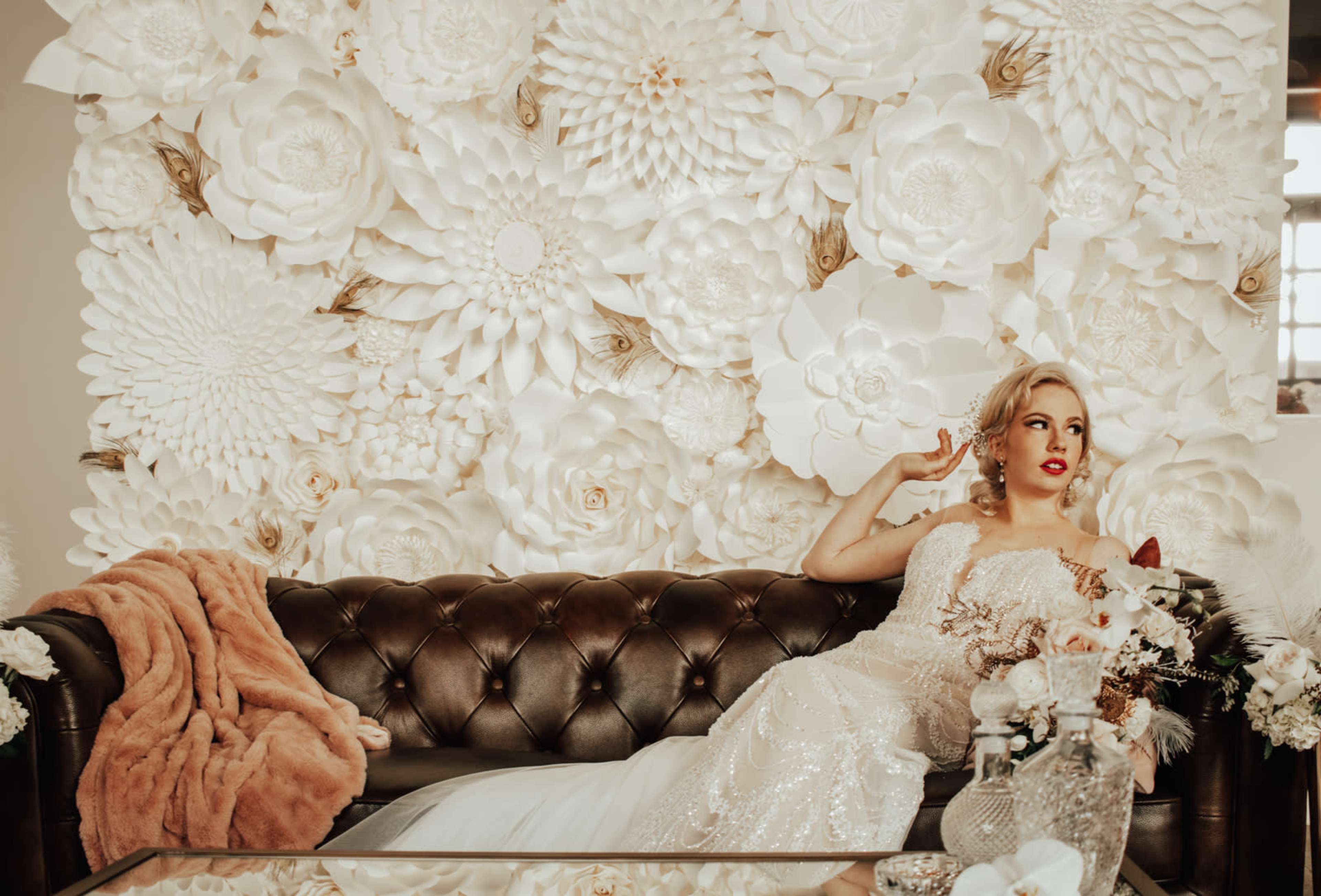 A bride in a sequined wedding gown poses on a dark leather sofa in front of a wall adorned with white paper flowers.