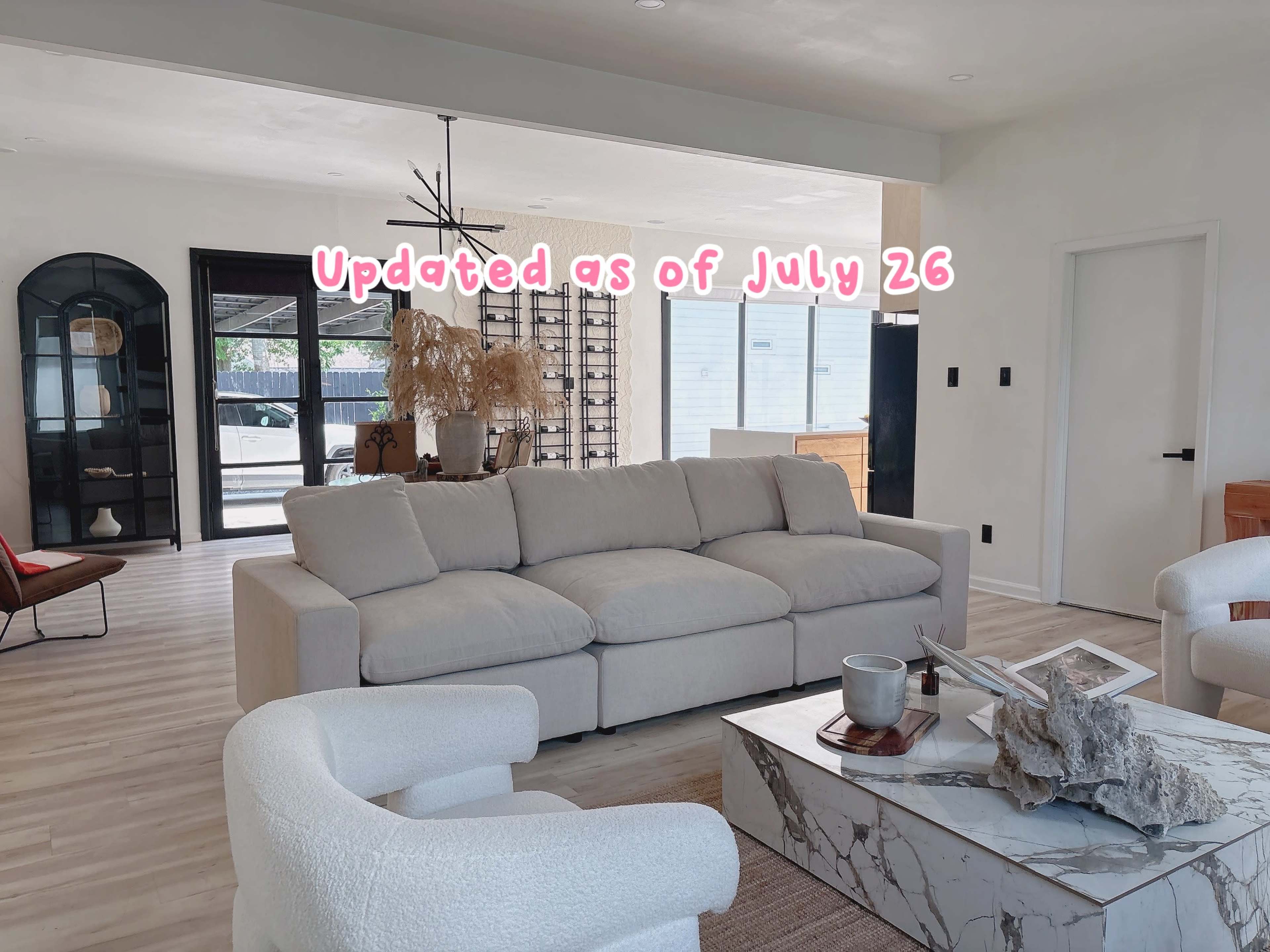 The image shows a modern living room featuring a light-colored sectional sofa, a marble coffee table, and several stylish chairs, with large windows allowing natural light to enter.
