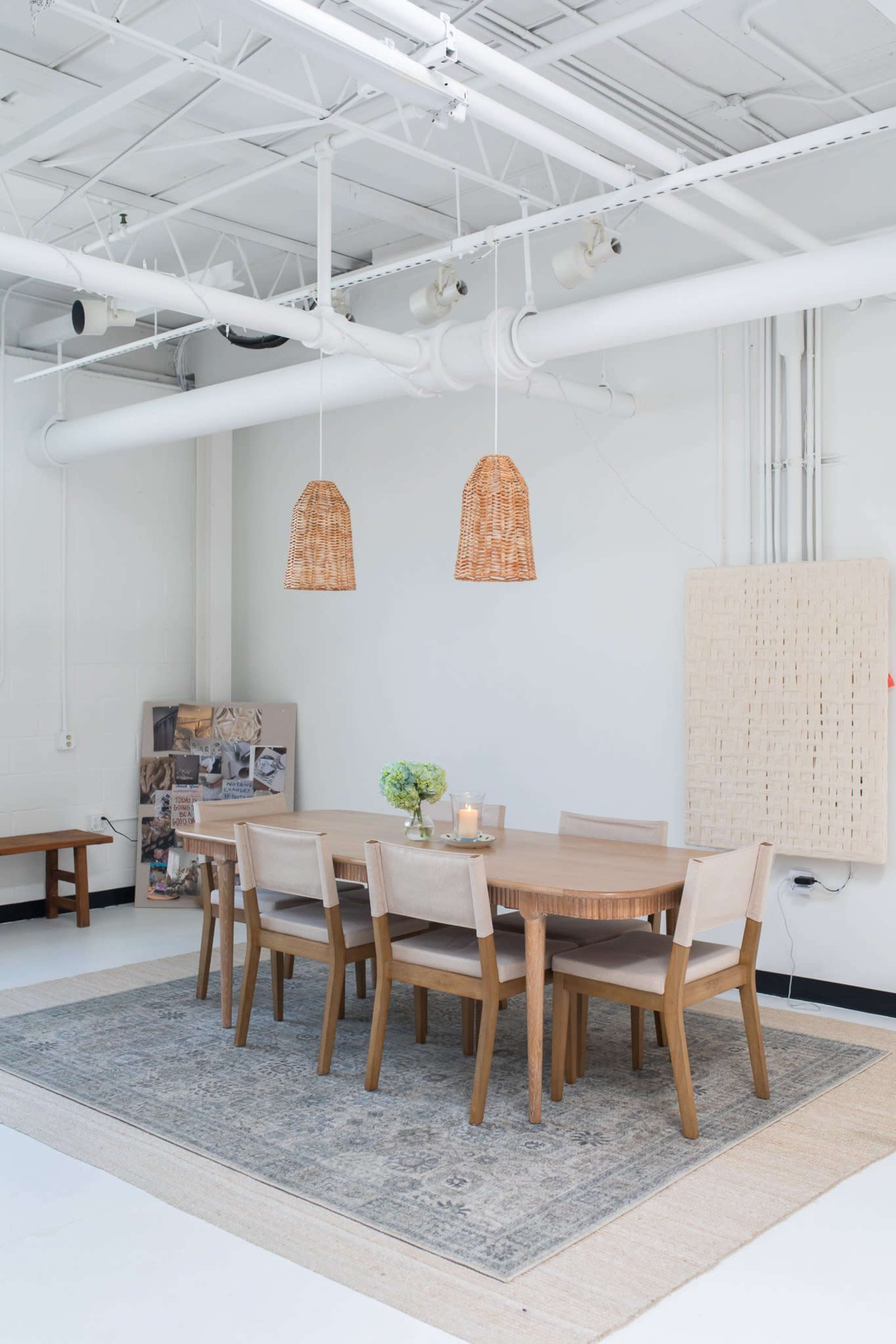 The image shows a modern dining area with a wooden table surrounded by six chairs, two woven pendant lights hanging above, and a decorative rug on the floor.