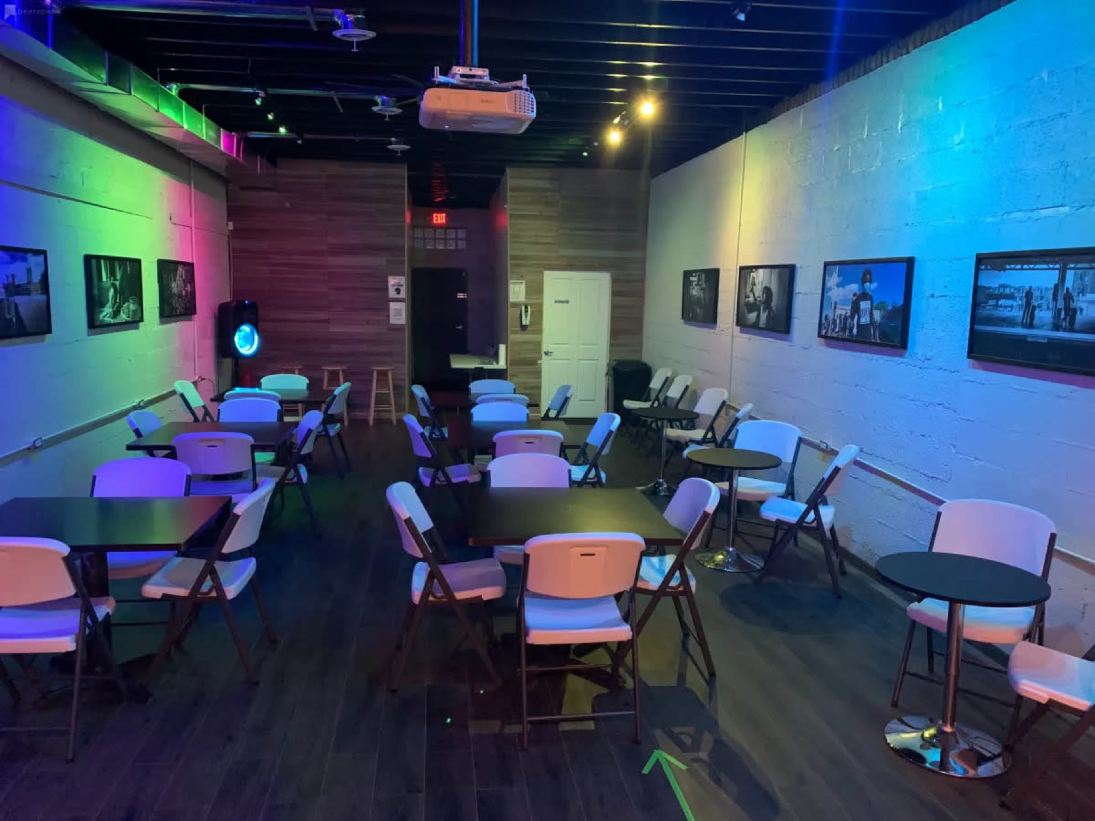 An empty event space with folding tables and chairs arranged, illuminated by colorful lights and featuring black-and-white photographs on the walls.