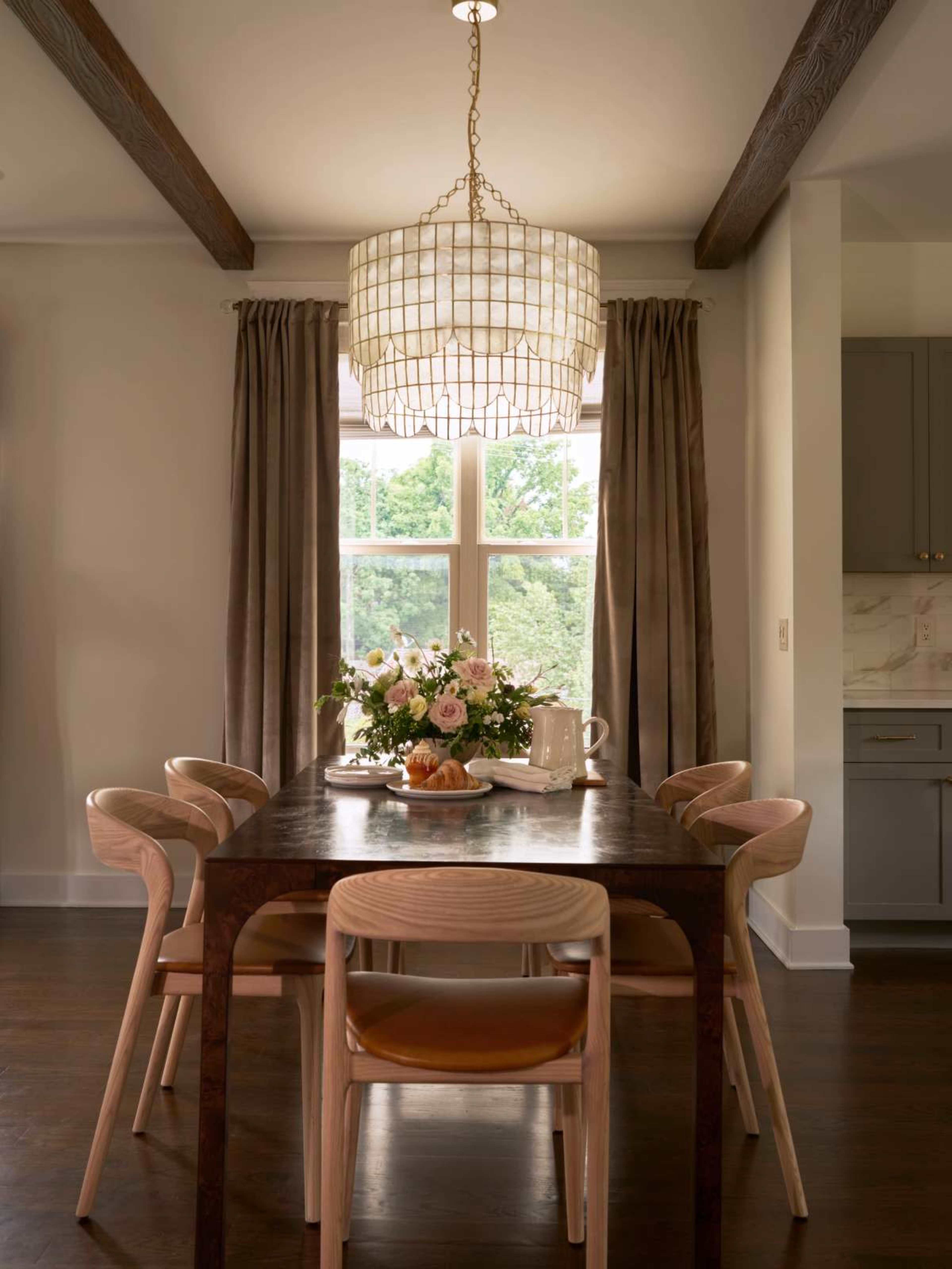 A dining room features a wooden table with four chairs, a vase of flowers, and a tea set, illuminated by a large pendant light above the table.