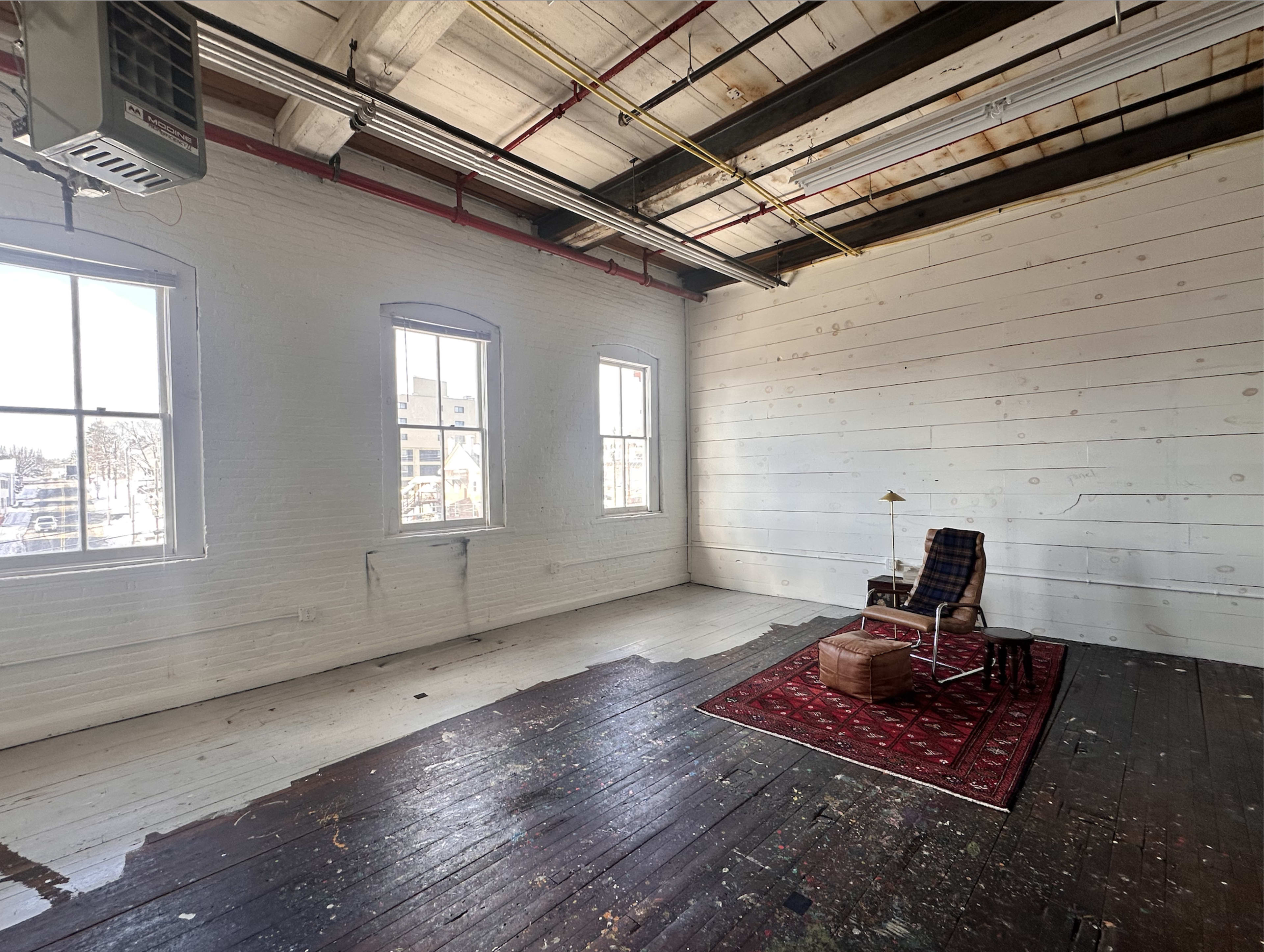 The image shows a spacious, empty room with exposed wooden beams, large windows, and a single chair on a patterned rug.
