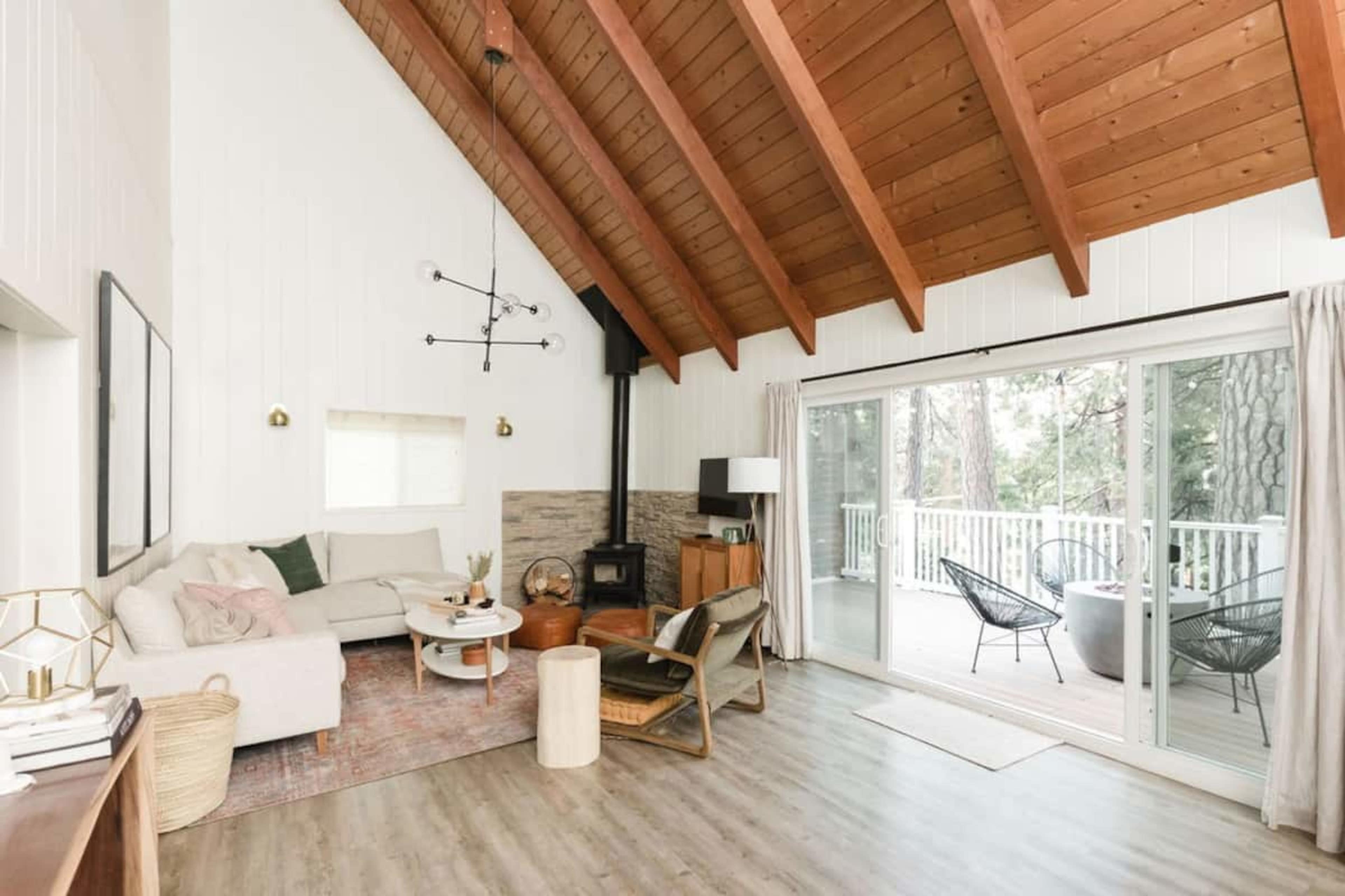 The image shows a modern living room with a high wooden ceiling, a white sofa, a coffee table, and large windows overlooking a deck with a seating area.