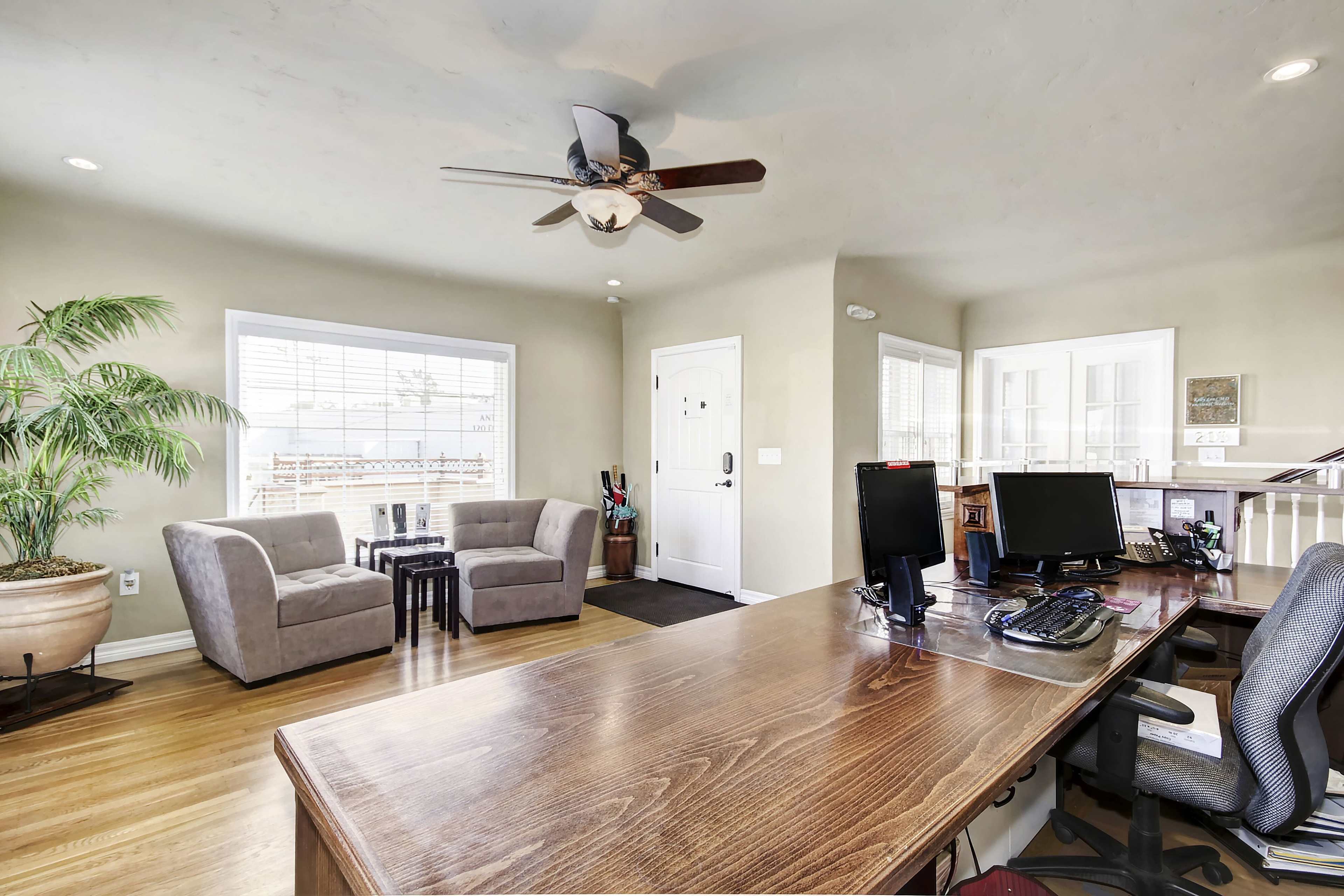 A home office with a wooden desk, two chairs, and a patio door, featuring a ceiling fan and a potted plant in the corner.