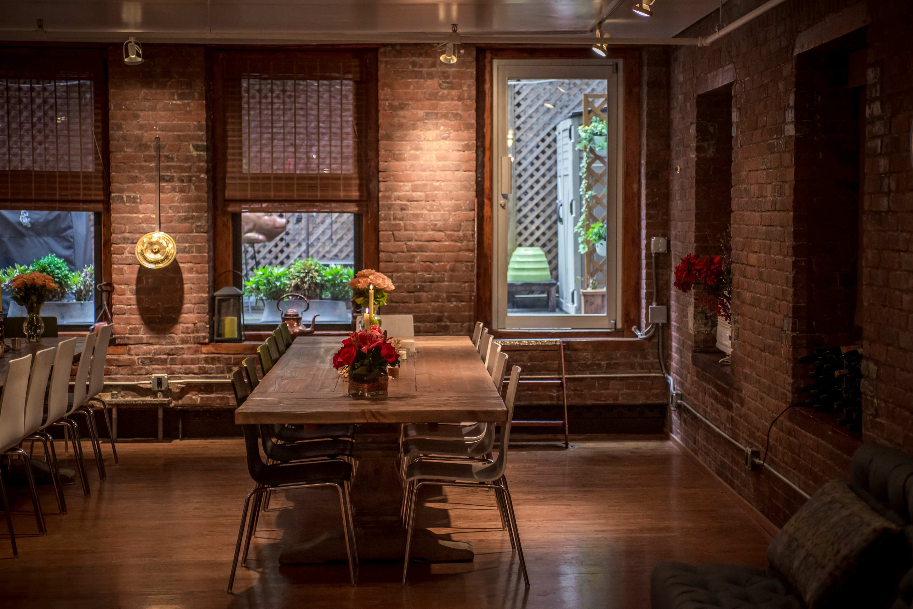 A long wooden dining table is set in a room with exposed brick walls and large windows that overlook a garden area.