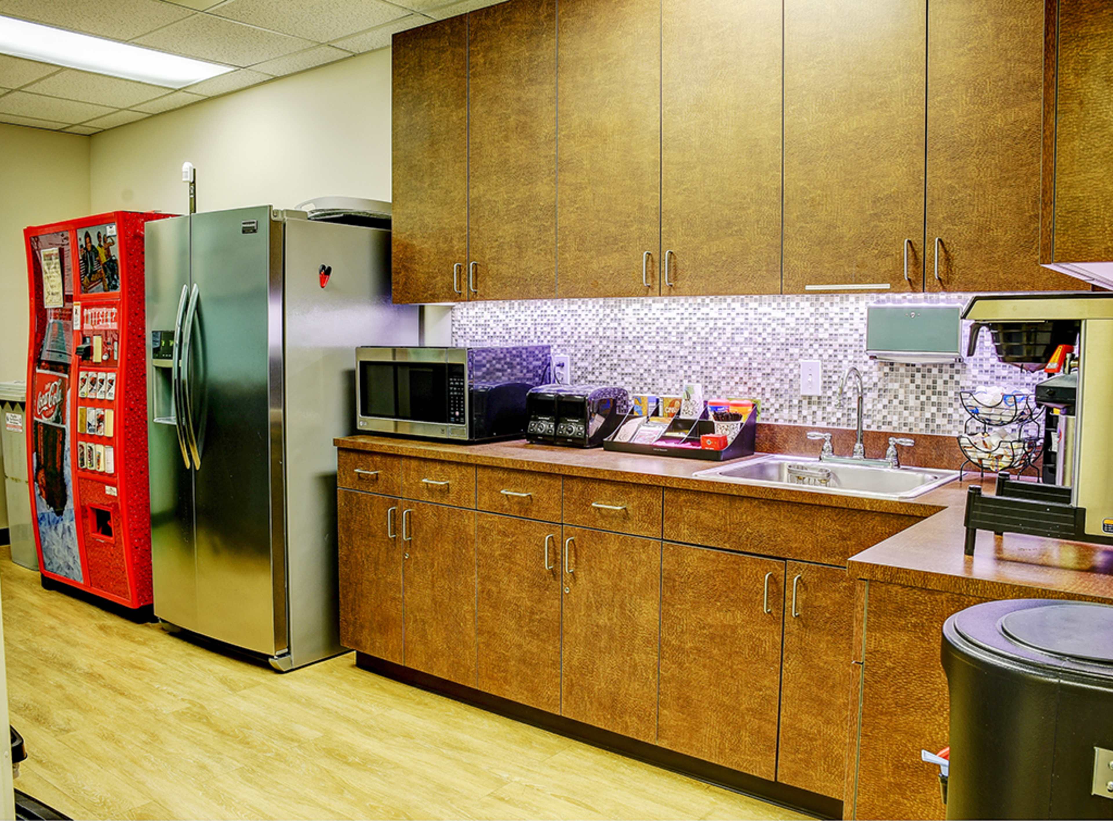 A kitchen area featuring a stainless steel refrigerator, a vending machine, a microwave, a coffee maker, and wooden cabinetry with a tiled backsplash.