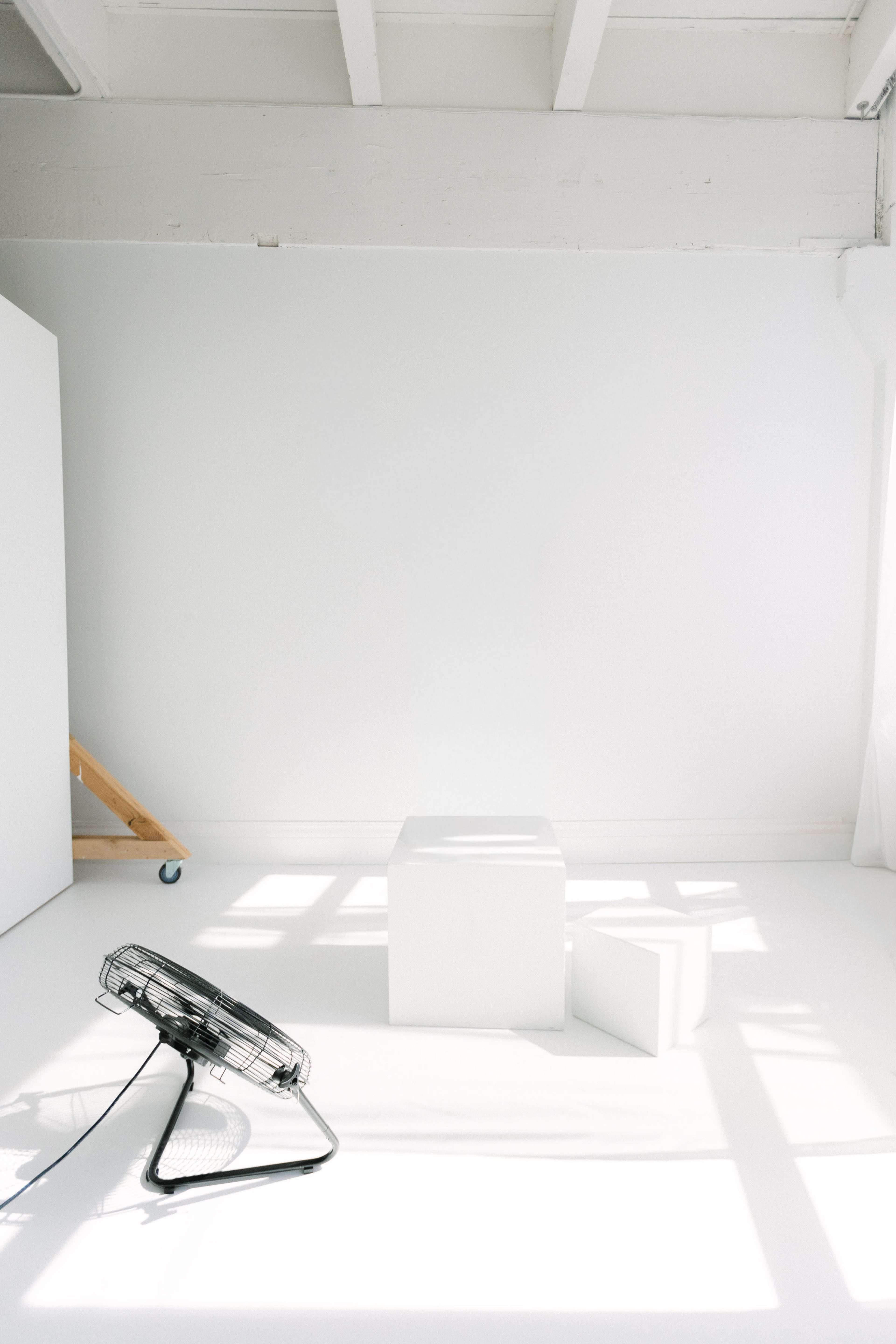The image shows a minimalistic room with a large white wall, two white cubes, and a black standing fan on a light-colored floor.