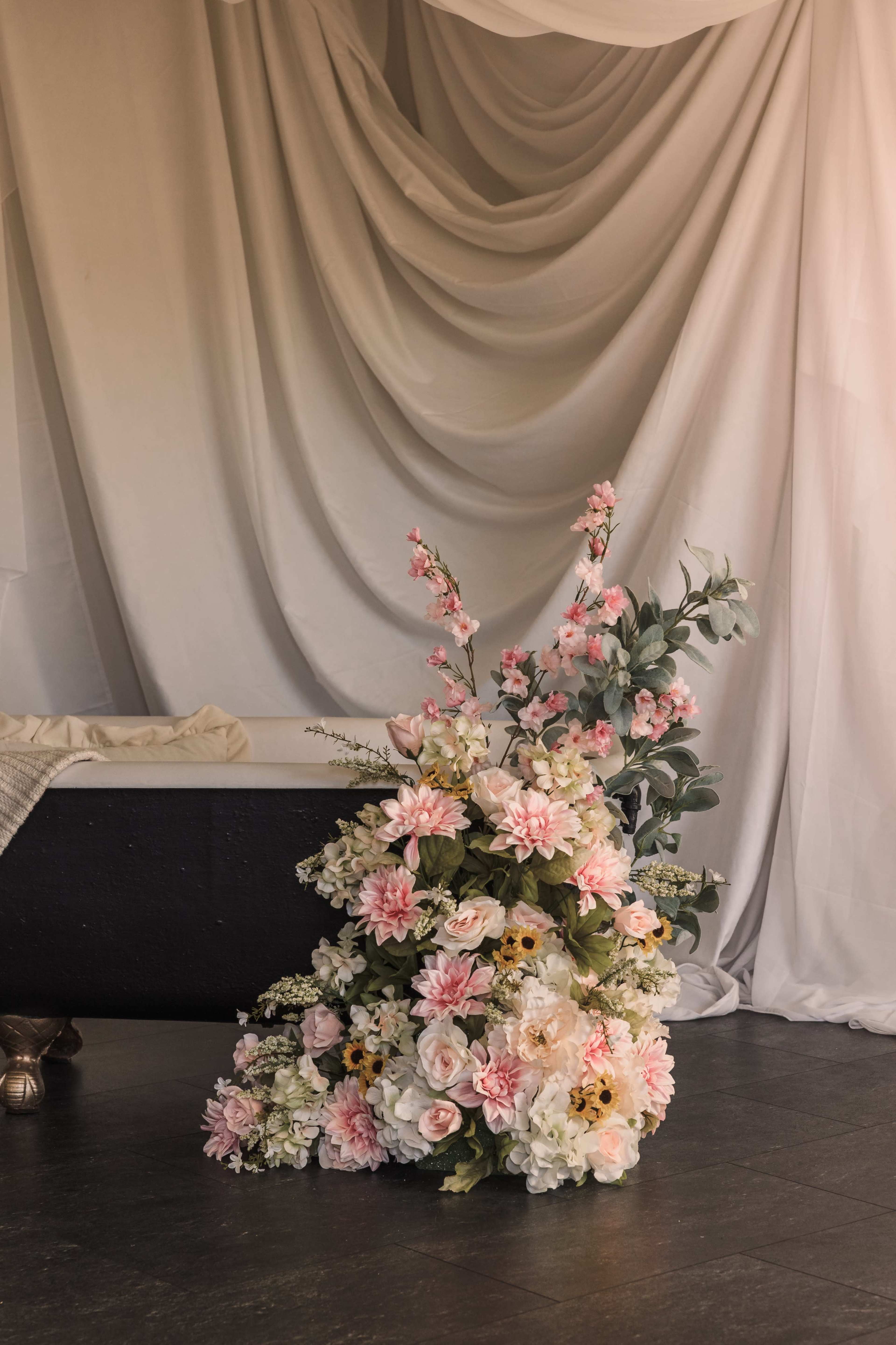 A large arrangement of artificial flowers in various shades of pink and white is placed on a dark floor, in front of a backdrop of softly draped white fabric.