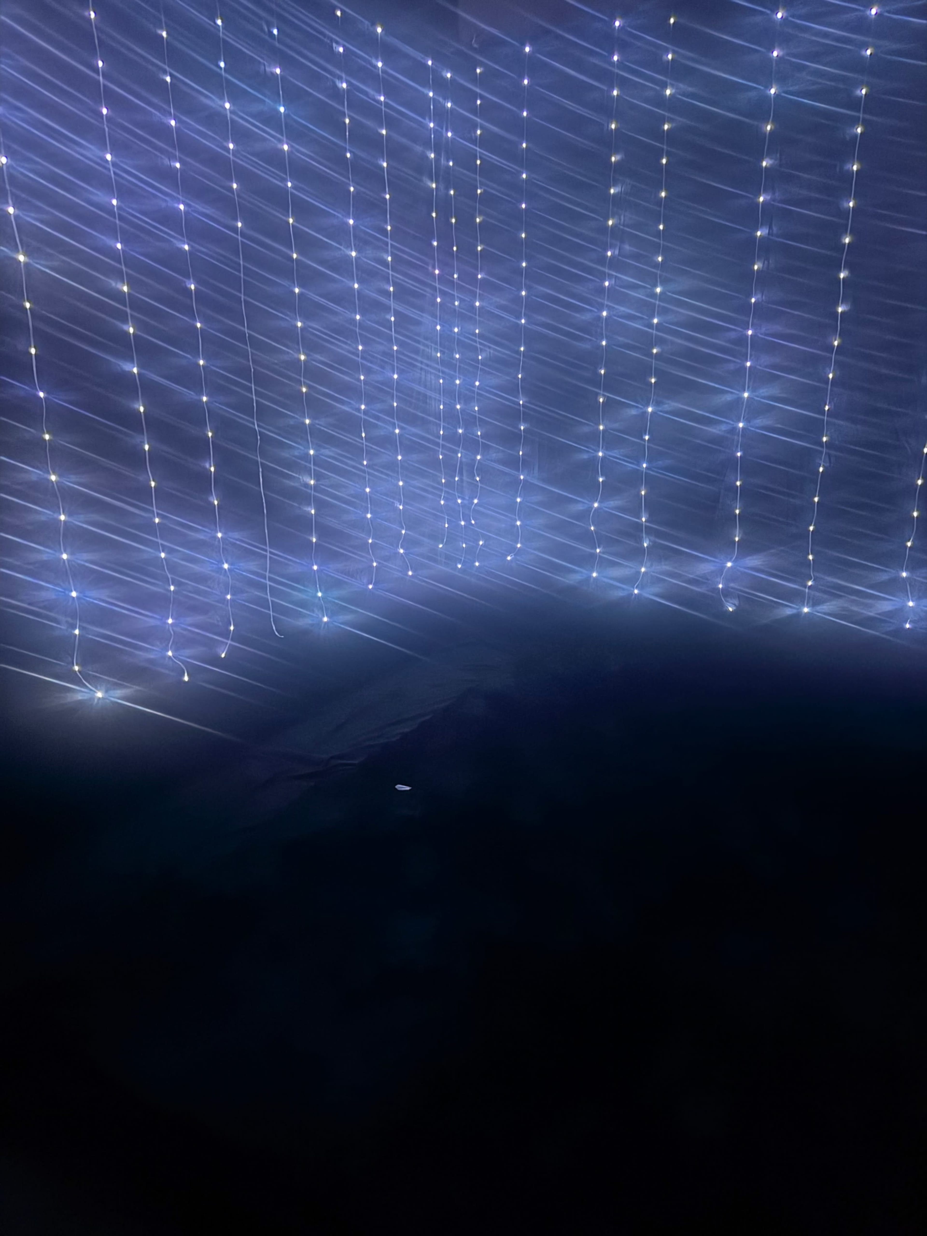 The image shows a dimly lit space with vertical strands of soft, colorful lights hanging in a cascading pattern.