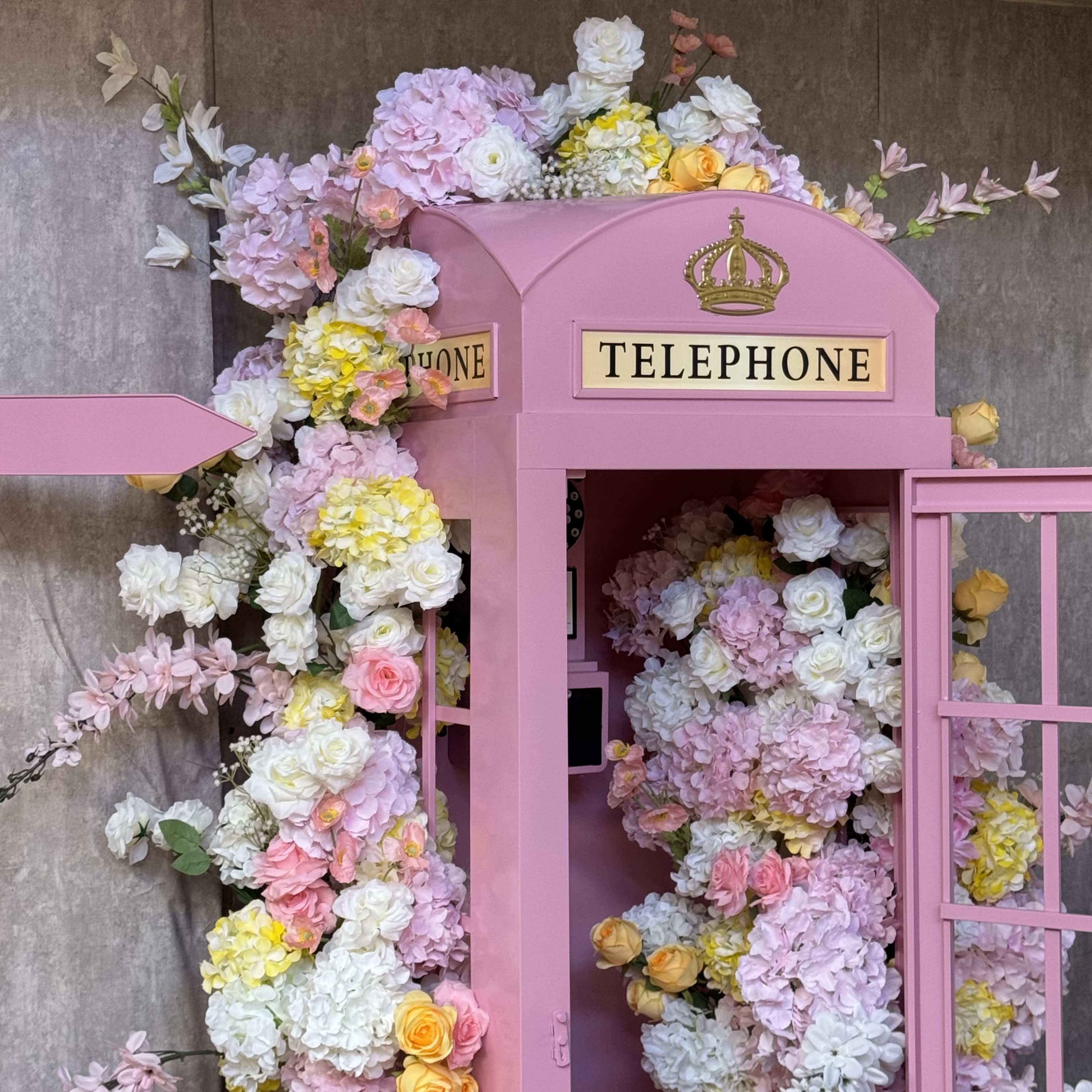 A pink telephone booth is adorned with a variety of colorful artificial flowers.