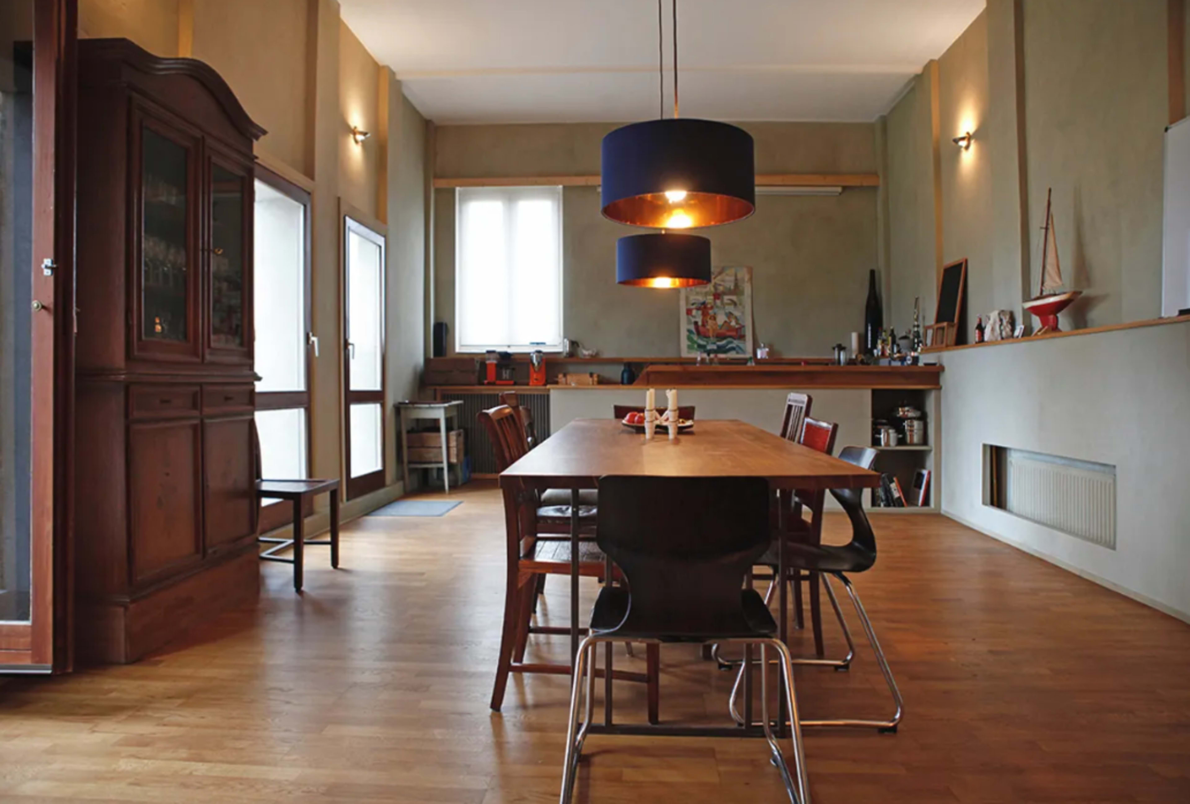 A spacious dining area features a long wooden table surrounded by various chairs, illuminated by a modern pendant light, with a cabinet and a bar area in the background.