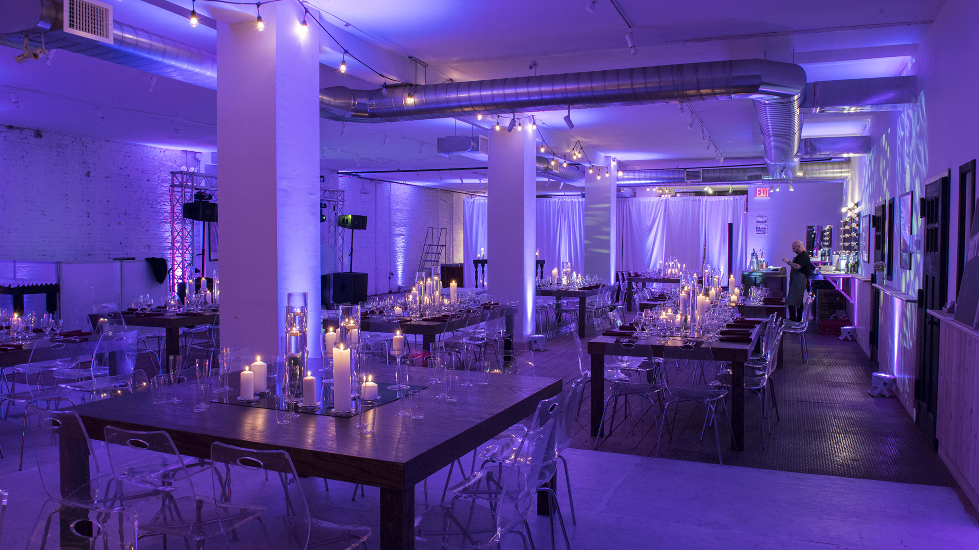 A spacious venue features several long wooden tables set for an event, illuminated by soft purple lighting and surrounded by candles and glass accents.