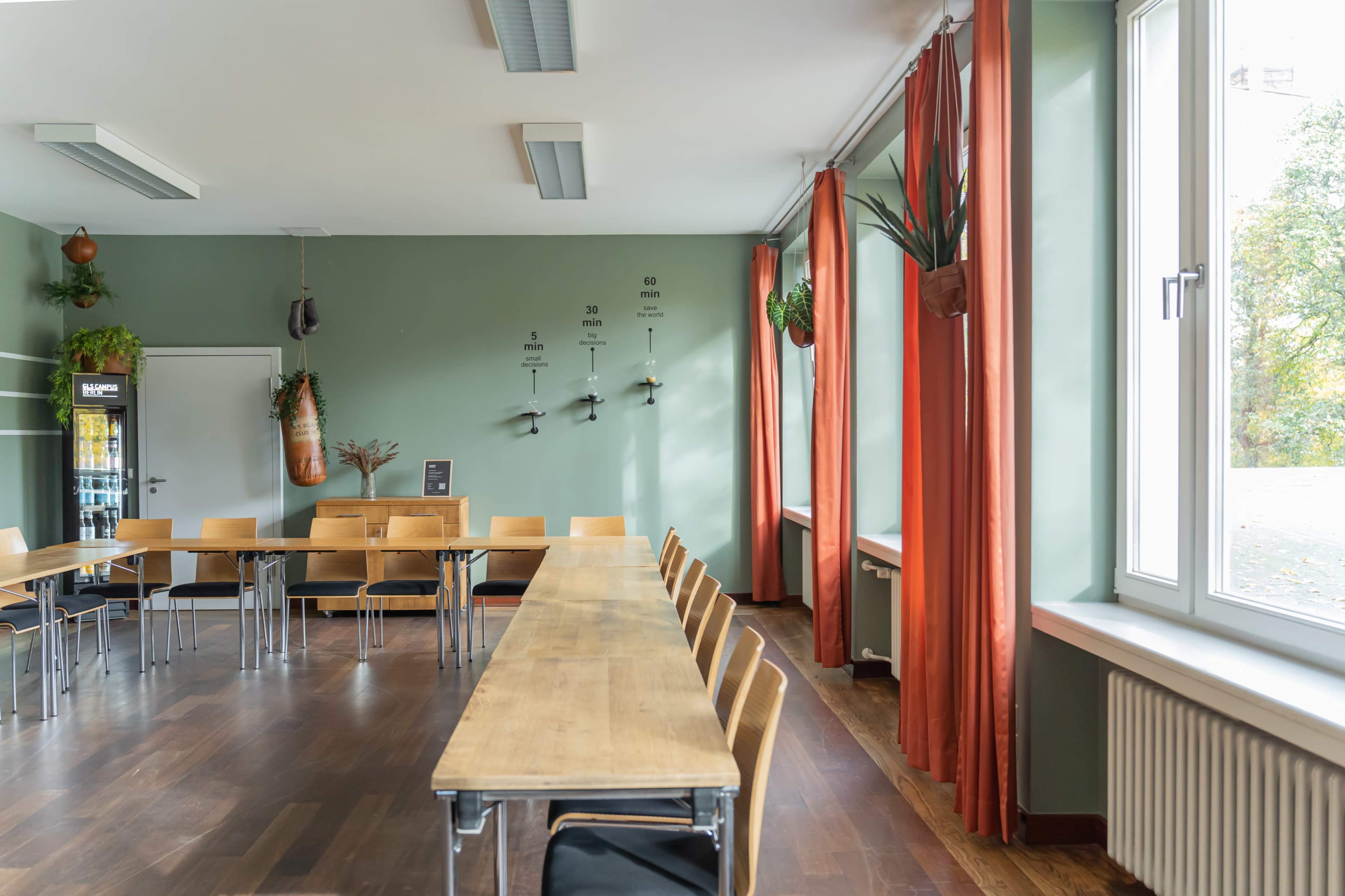 The image shows a meeting room with wooden tables arranged in a U-shape, surrounded by chairs, and large windows adorned with orange curtains.