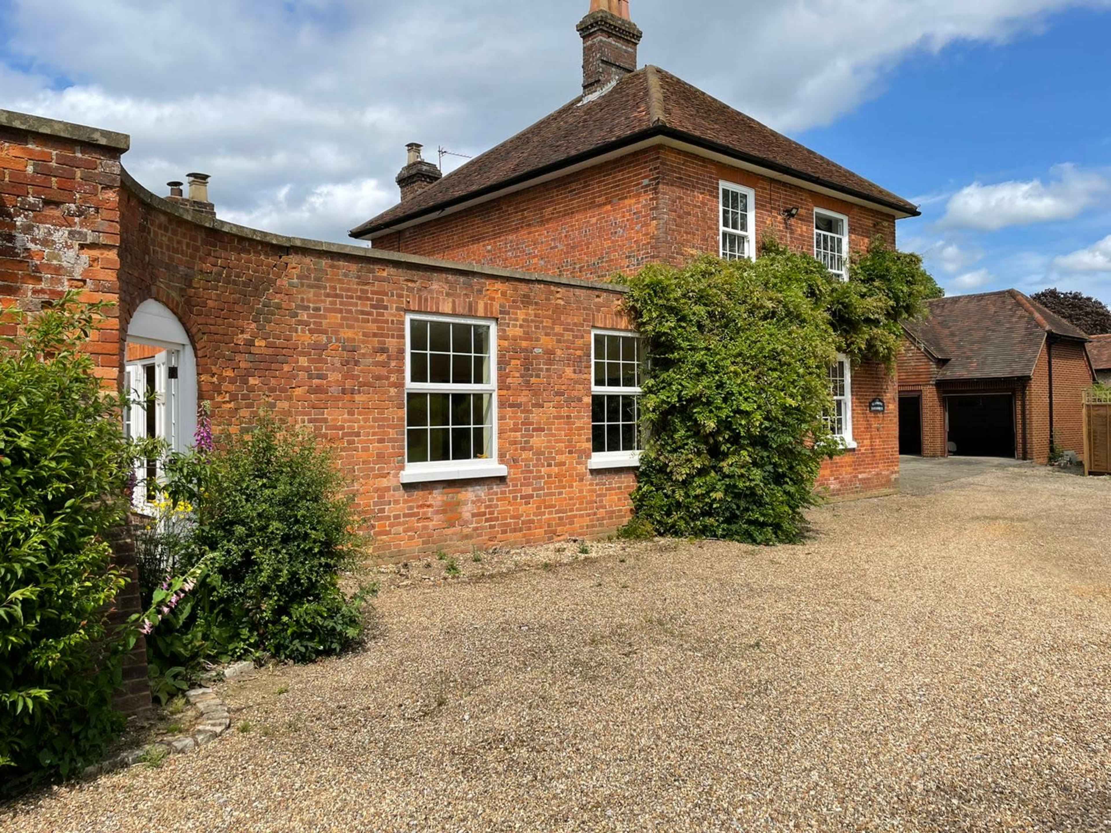A two-story brick house with white window frames is situated beside a gravel driveway and is partially covered by green vegetation.