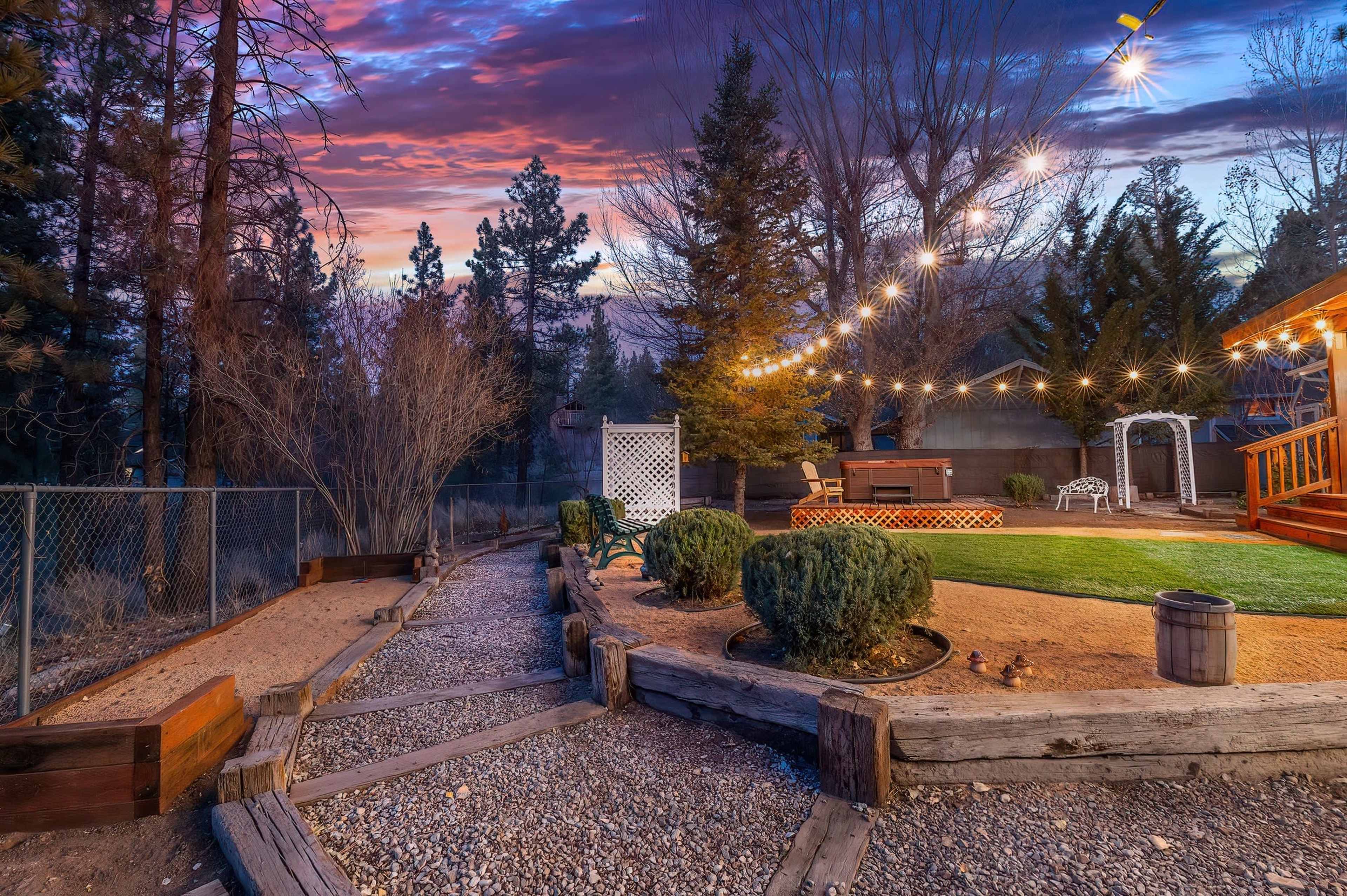 A well-lit outdoor patio area features a path lined with gravel, leading to a landscaped yard with trees and string lights against a colorful sunset sky.