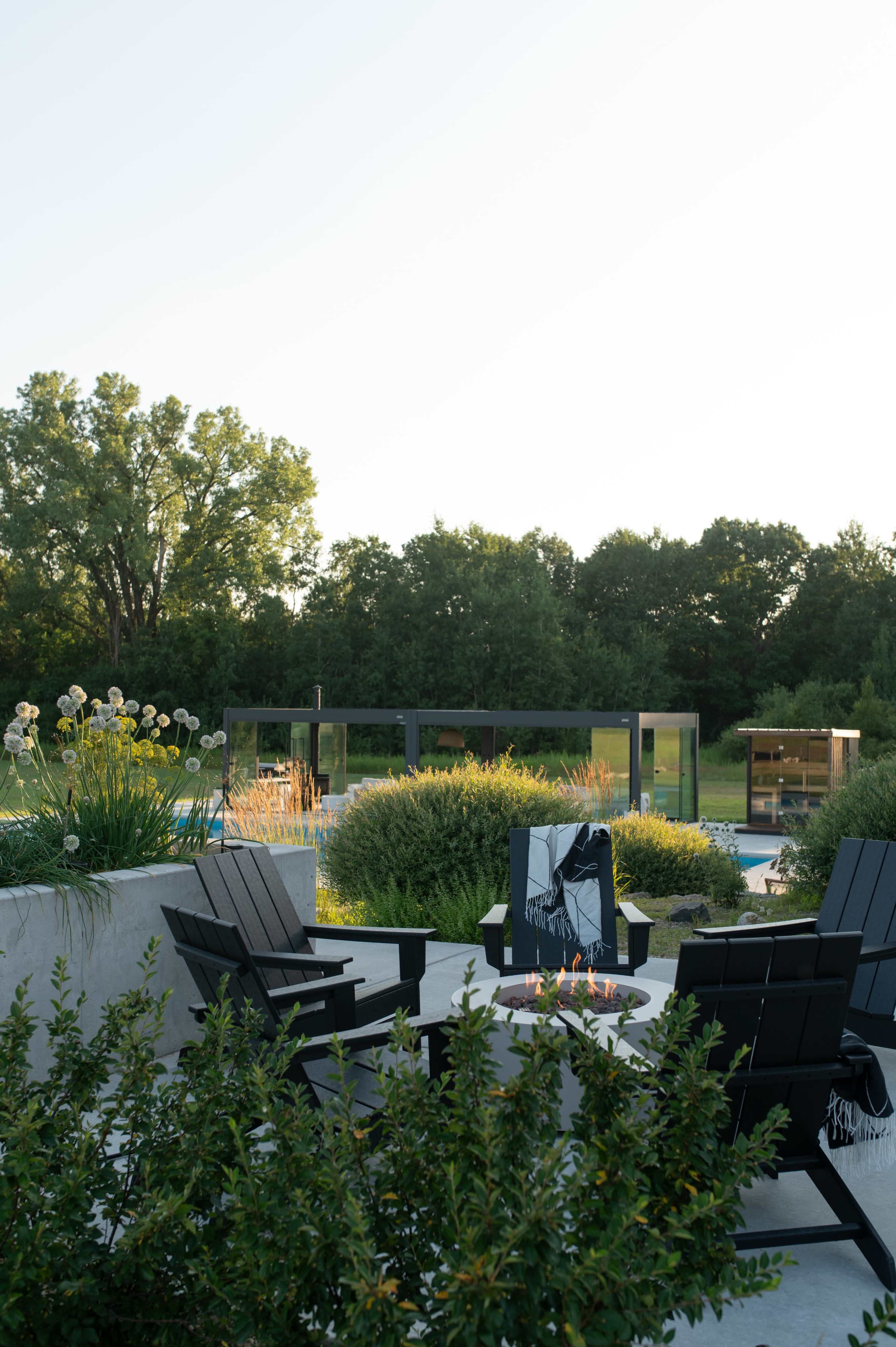 The image shows a modern outdoor seating area with black chairs surrounding a fire pit, set against a backdrop of greenery and a swimming pool.
