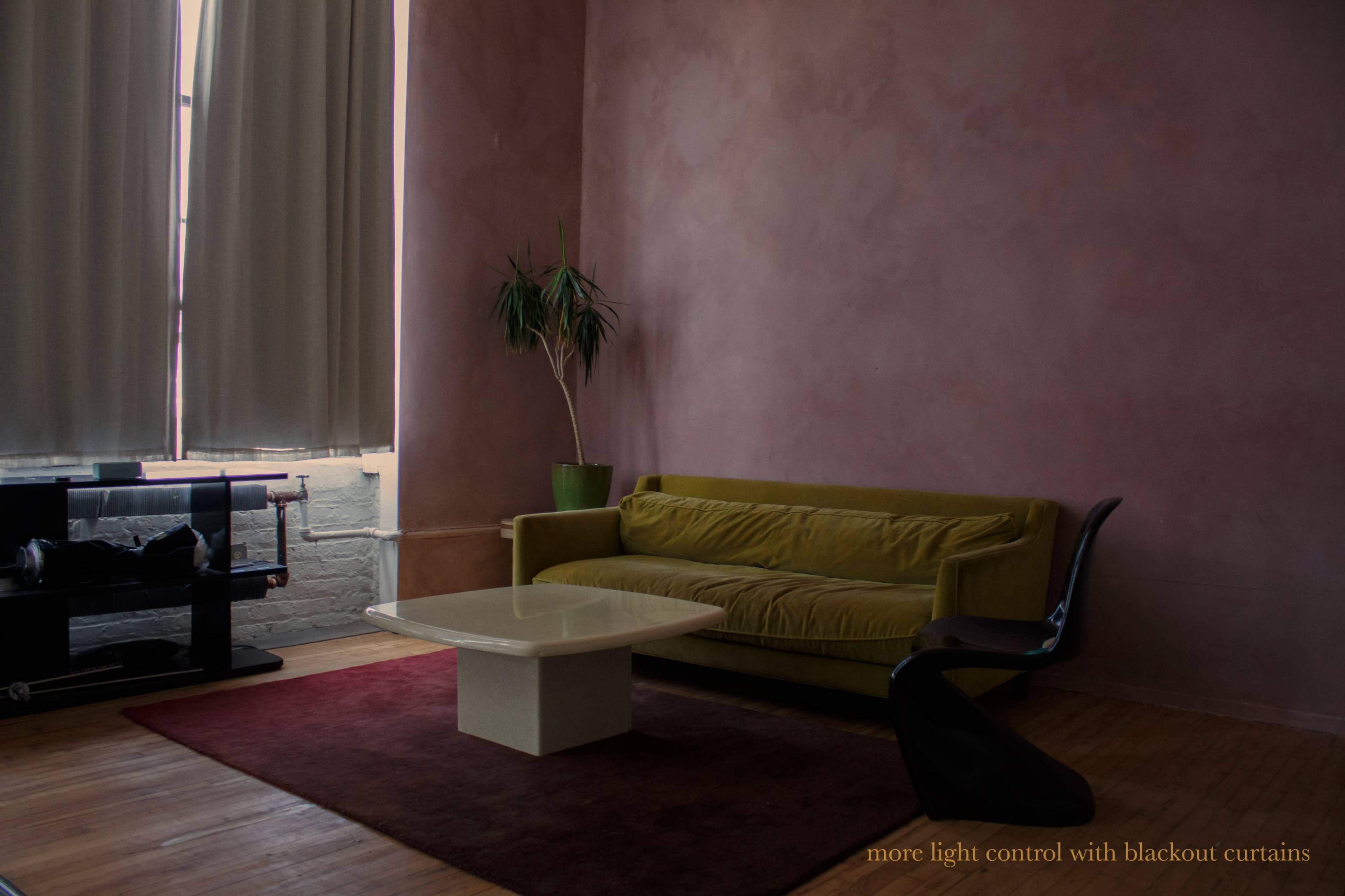 A green couch sits in a room with a pink wall, accompanied by a light-colored coffee table and a tall plant next to a window covered with long curtains.