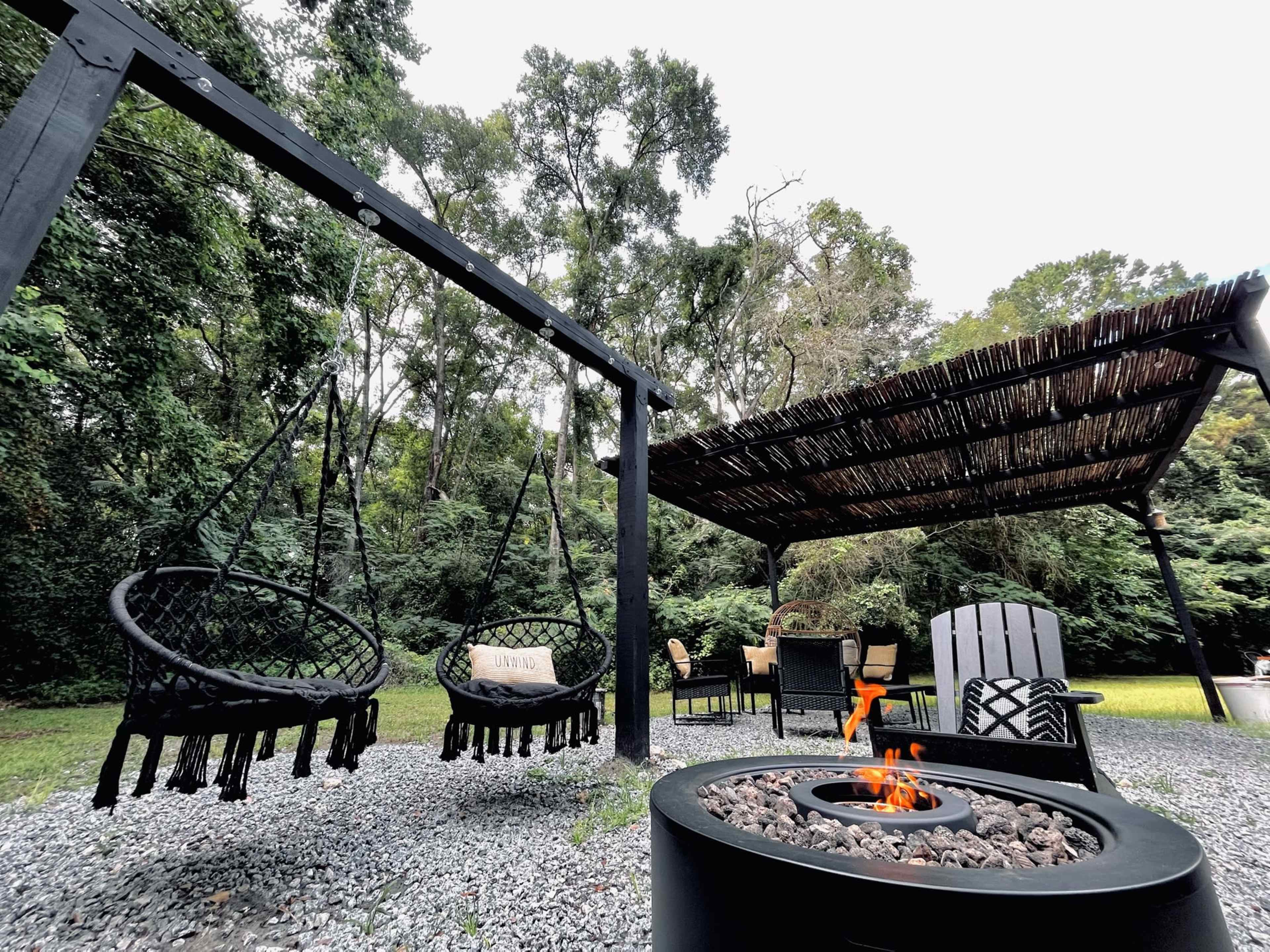 A outdoor patio area with two hanging chairs, a fire pit, and a covered seating space surrounded by trees.