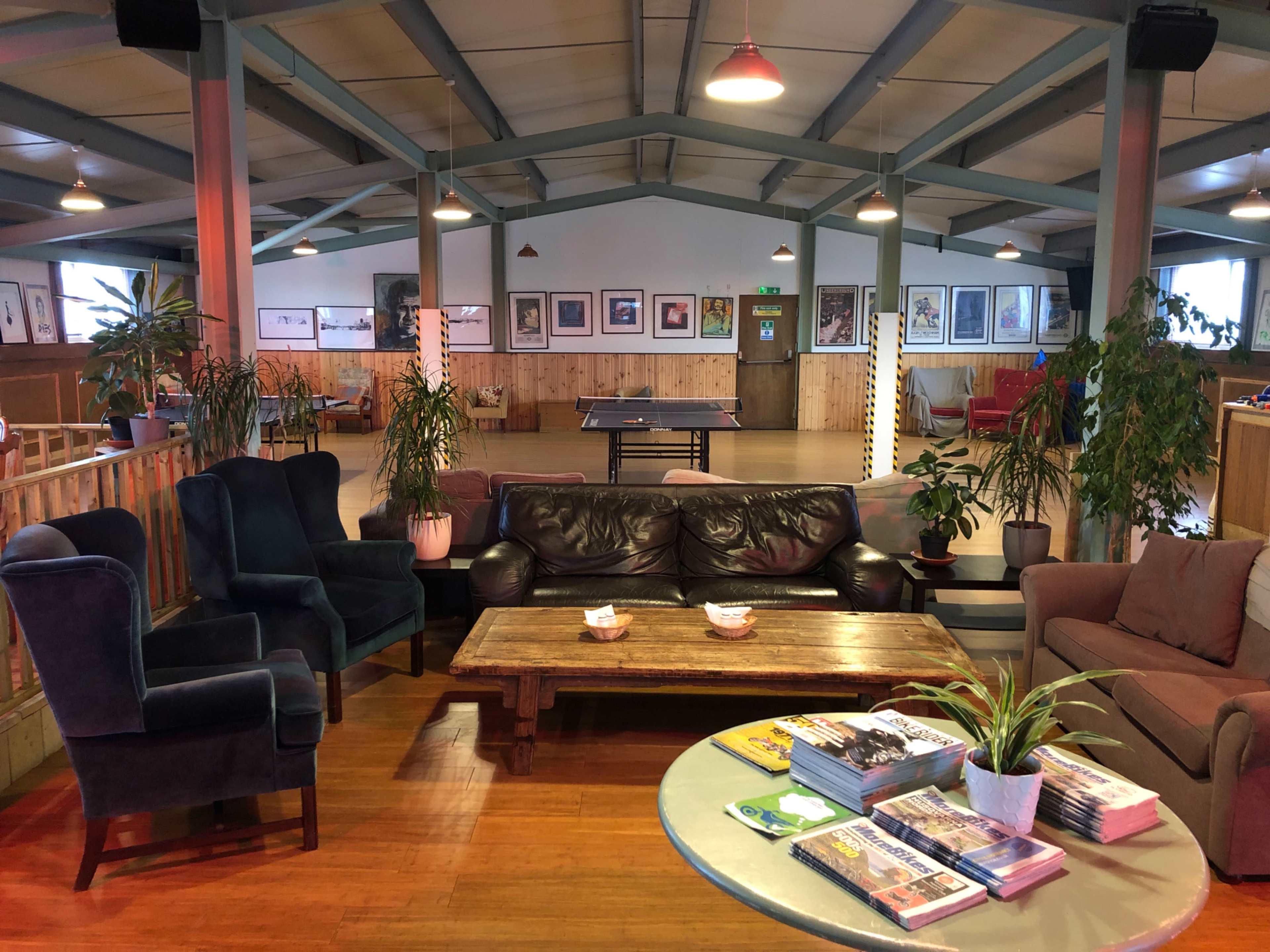 A spacious, well-lit indoor lounge area with seating, plants, a coffee table, and a ping-pong table in the background.