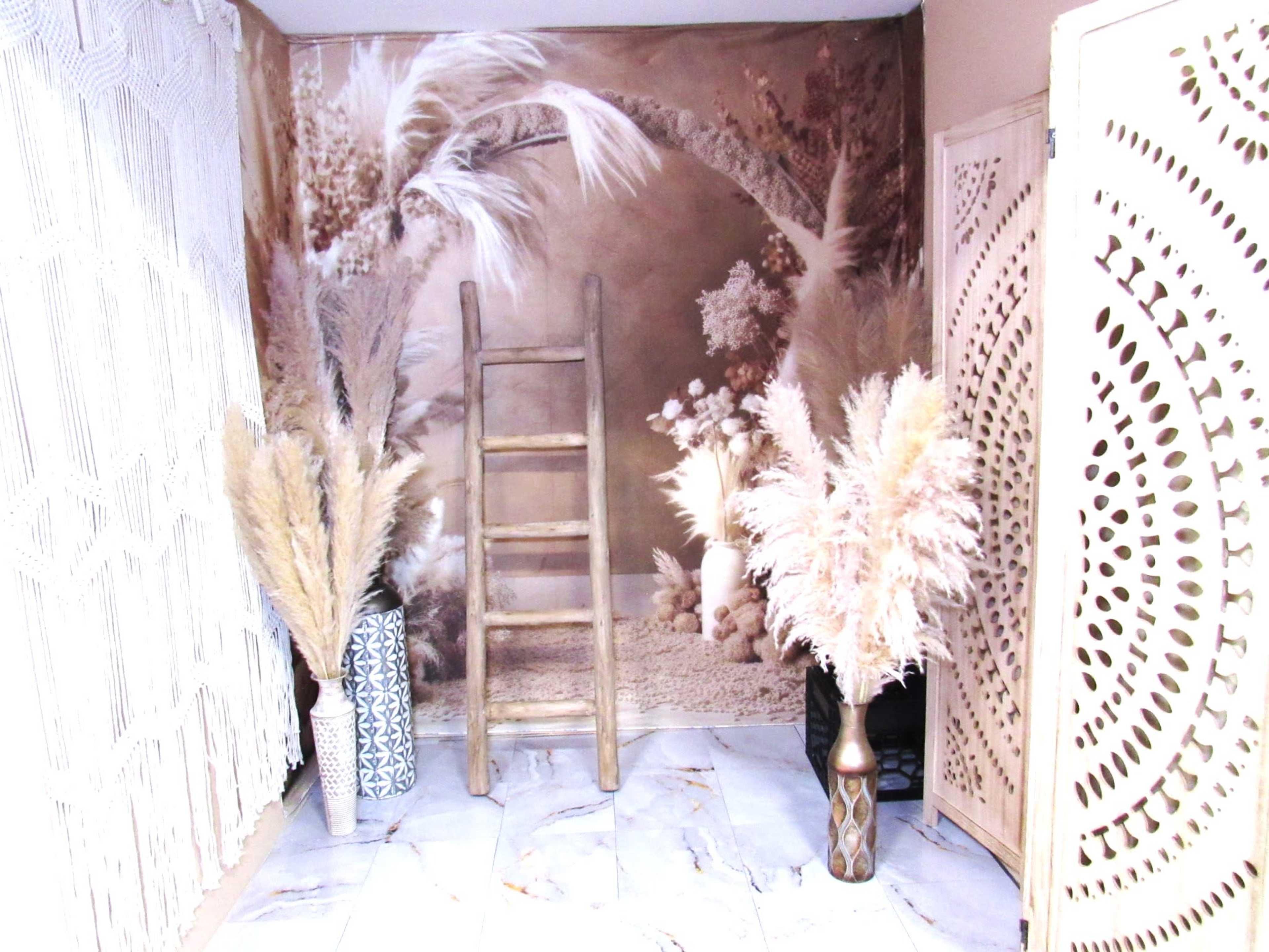 A room features a ladder against a backdrop of decorative plants, with large pampas grass arrangements in vases on either side.
