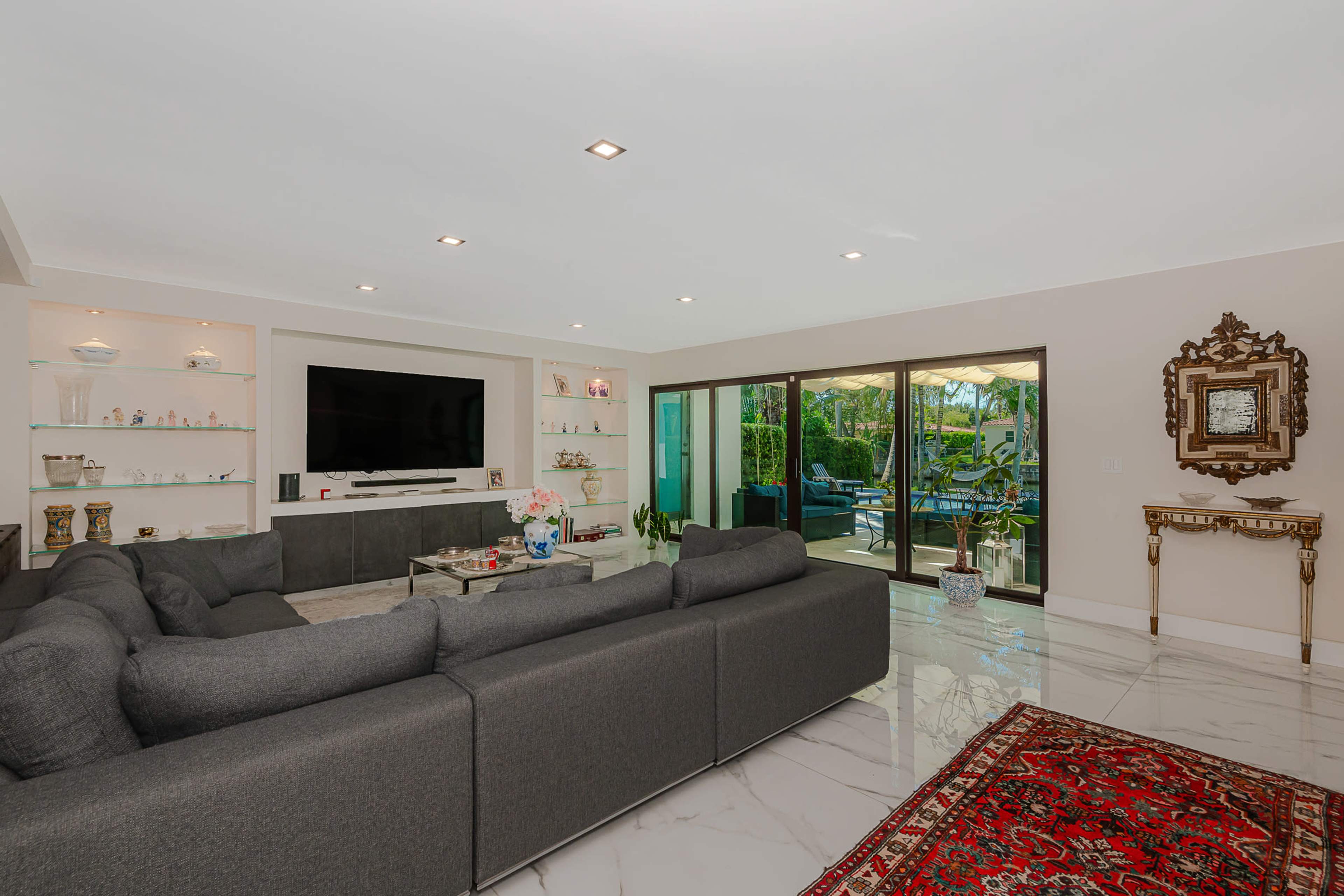 The image shows a modern living room with a large gray sectional sofa, a mounted television on the wall, and sliding glass doors leading to an outdoor patio.