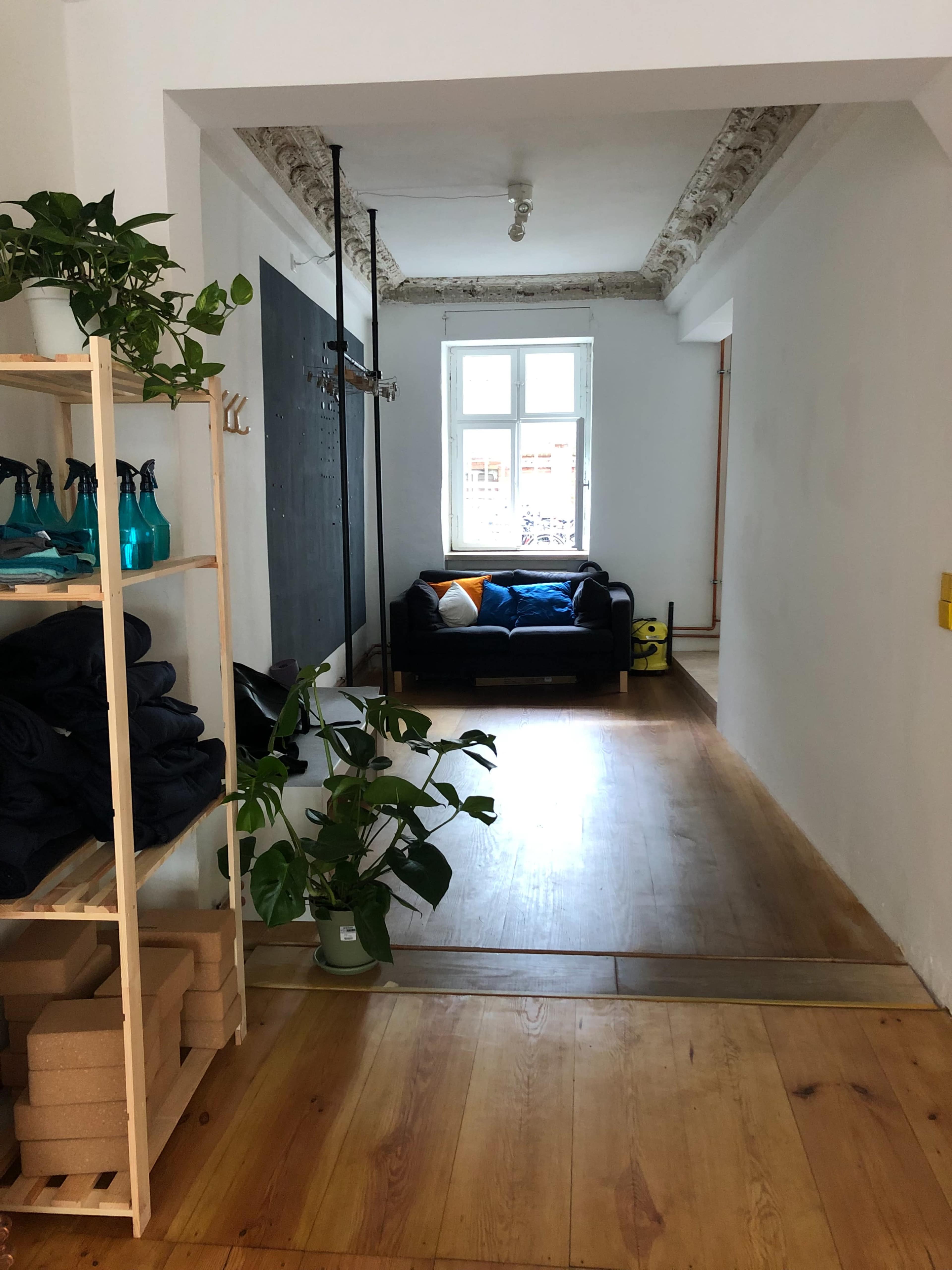 The image shows a bright, minimalist interior space featuring a wooden floor, plants, a couch, and a large window.