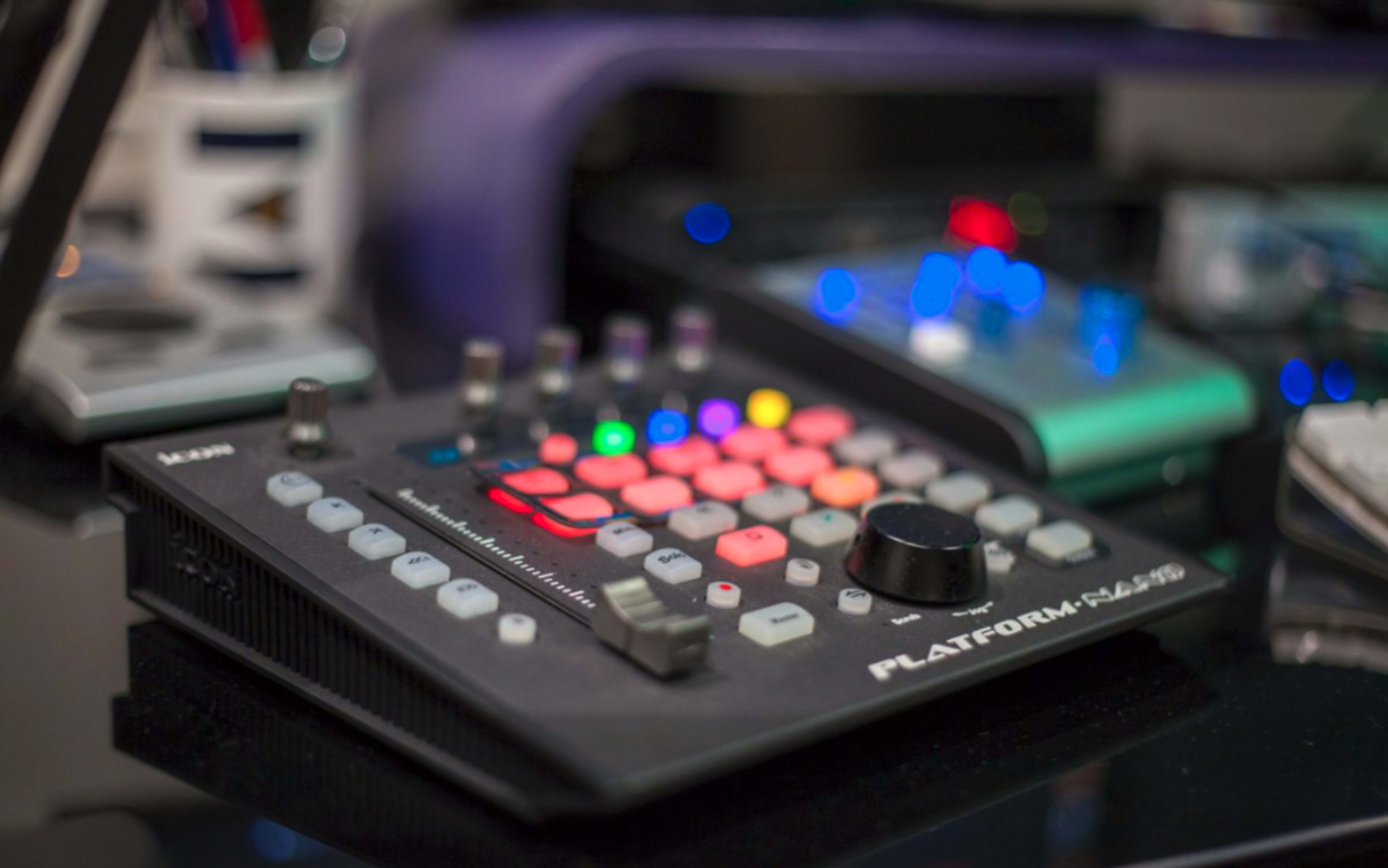 A MIDI controller with colorful buttons and a large knob sits on a desk among other electronic devices.