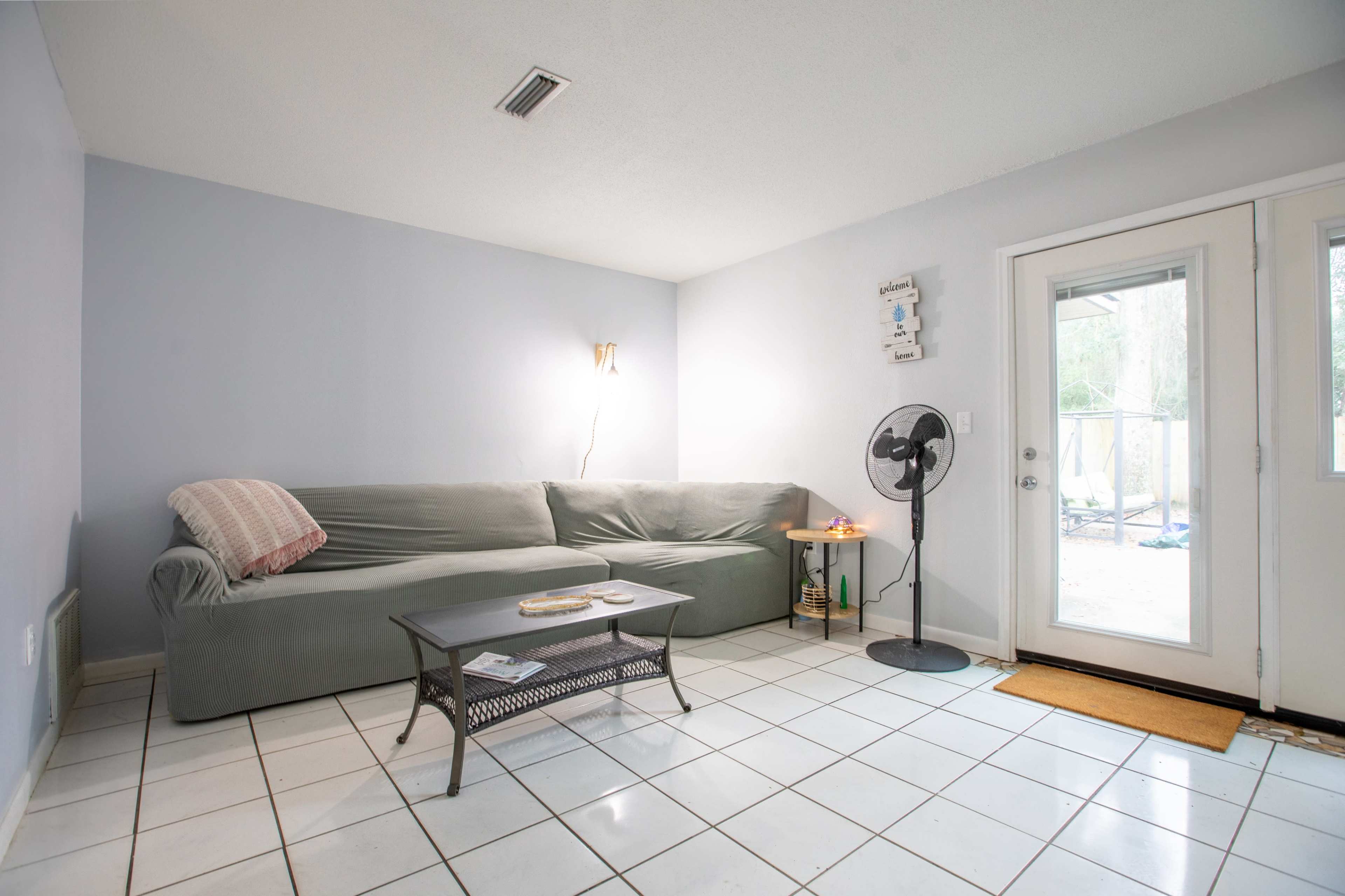 A light blue living room features a green sofa, a small table, a standing fan, and a door leading outside.