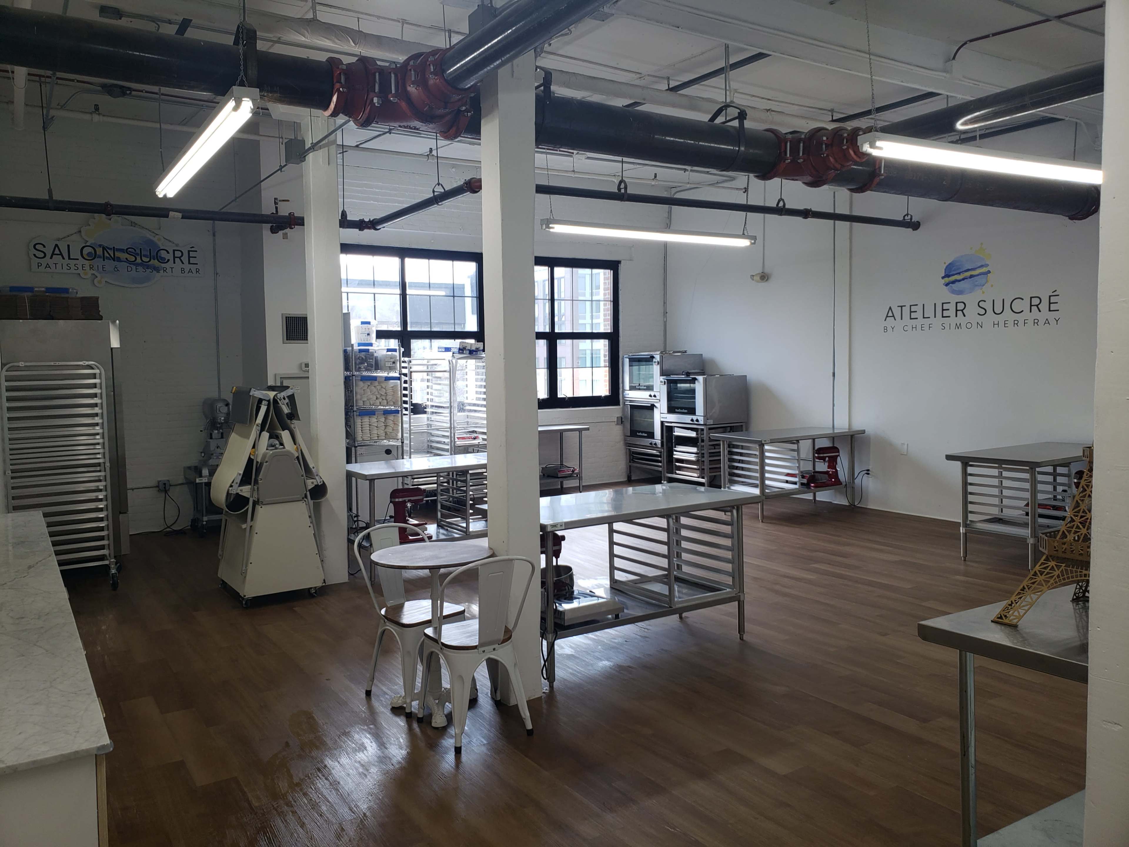 The image shows a spacious and well-lit kitchen area with stainless steel tables, baking equipment, and shelves, featuring branding for "Atelier Sucré" by Chef Simon Herrray on the wall.