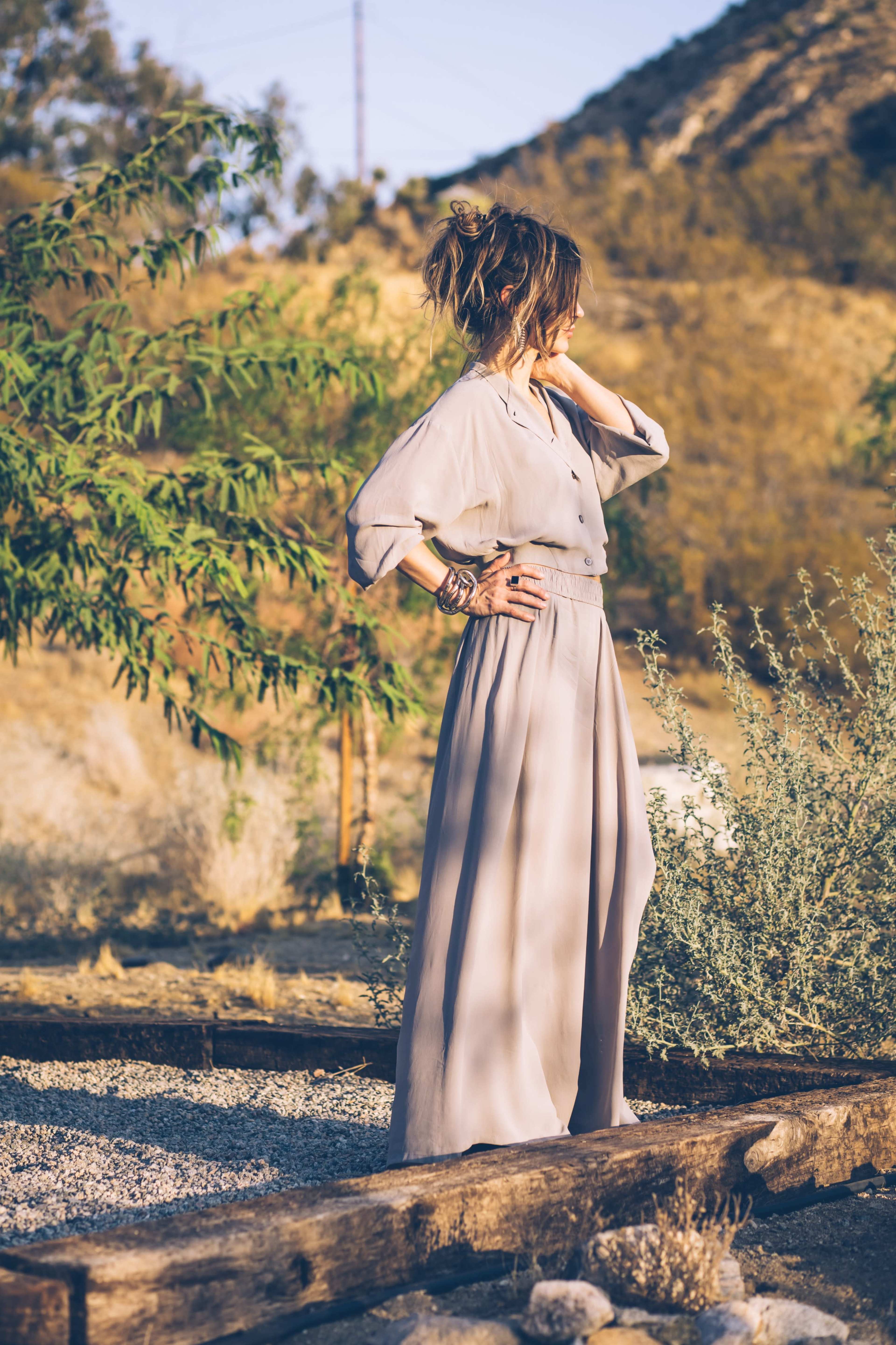A woman stands in a flowy outfit next to sparse vegetation in a rocky outdoor setting.