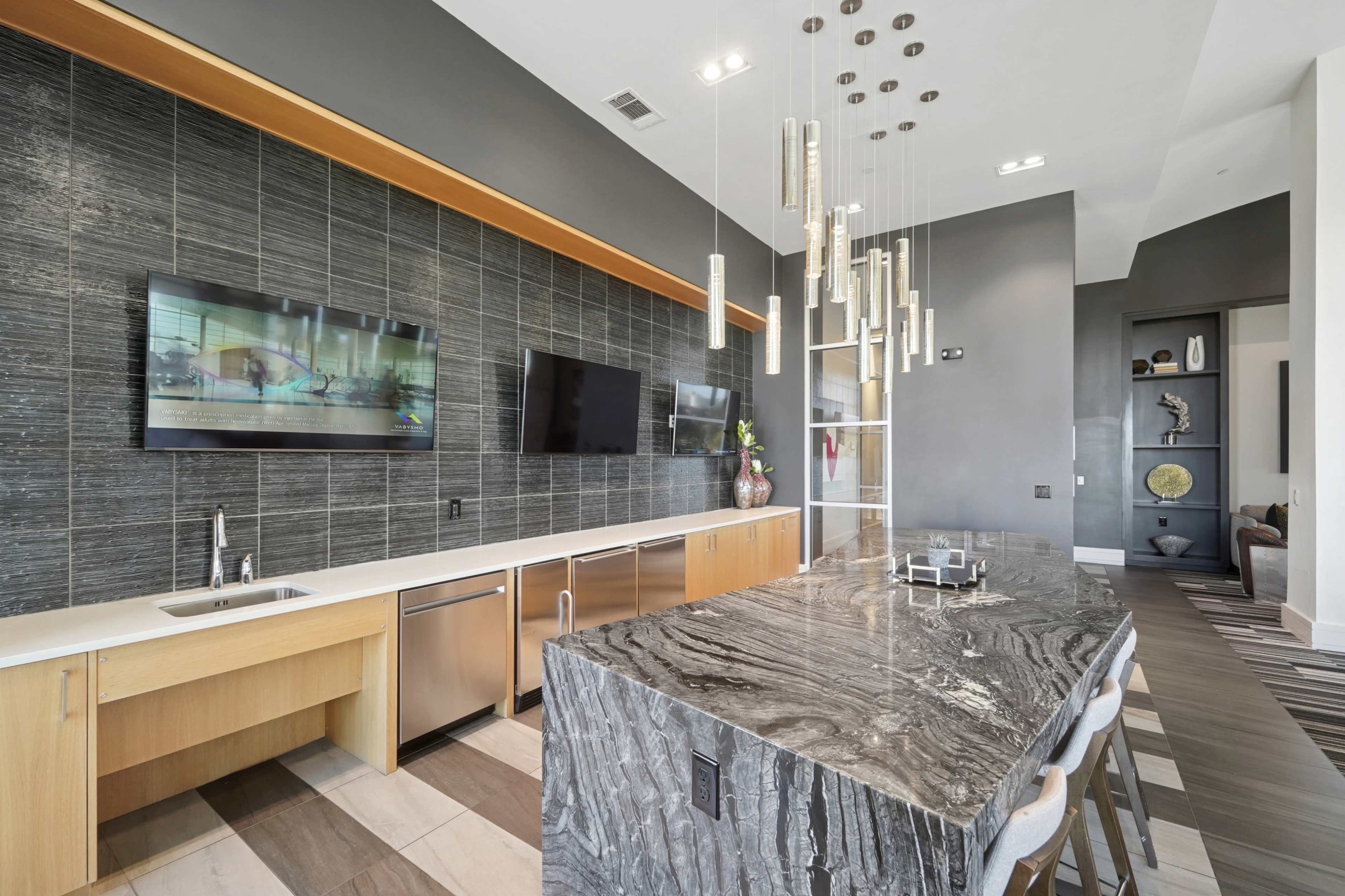 The image shows a modern kitchen featuring a large island with a textured surface, multiple pendant lights overhead, and sleek cabinetry against a dark, tiled wall.