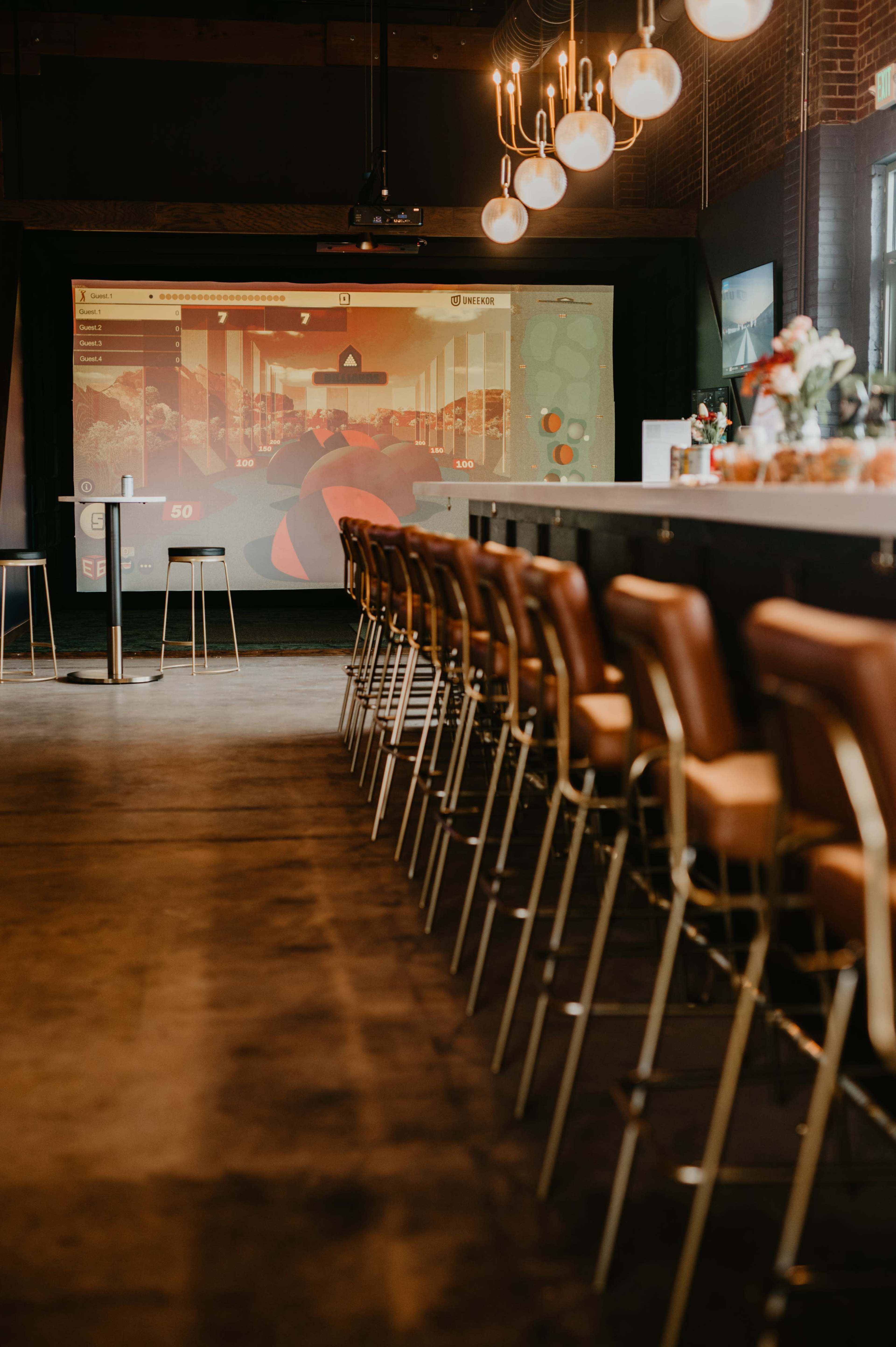 A modern bar with brown leather stools lined up in front of a large screen displaying a colorful digital interface.