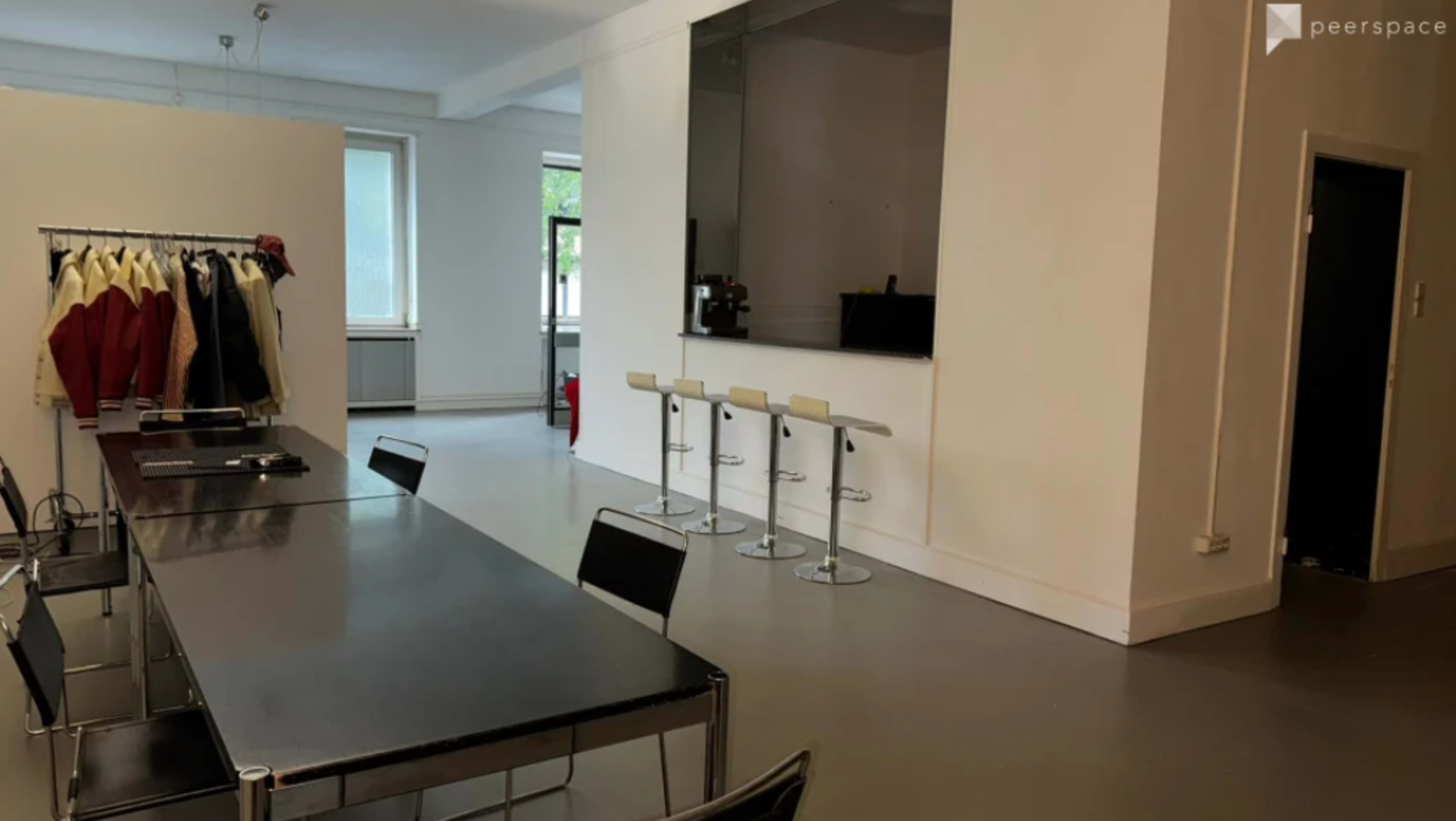 The image shows a modern interior space with a long table, several black chairs, a clothing rack, and a bar area with three high stools.