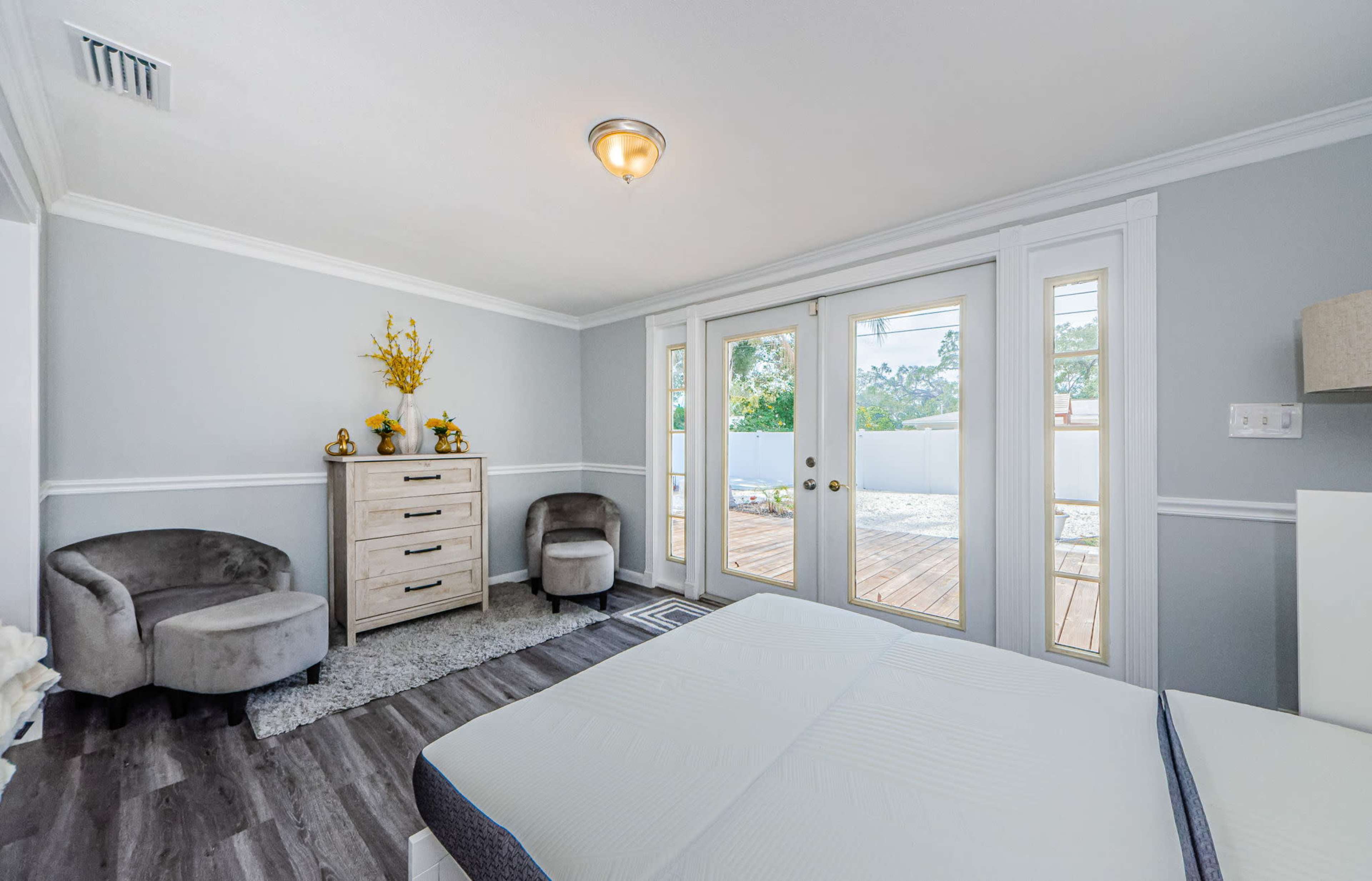 A bedroom featuring a light gray color scheme, a dresser, two chairs, and sliding glass doors leading to a wooden deck outside.